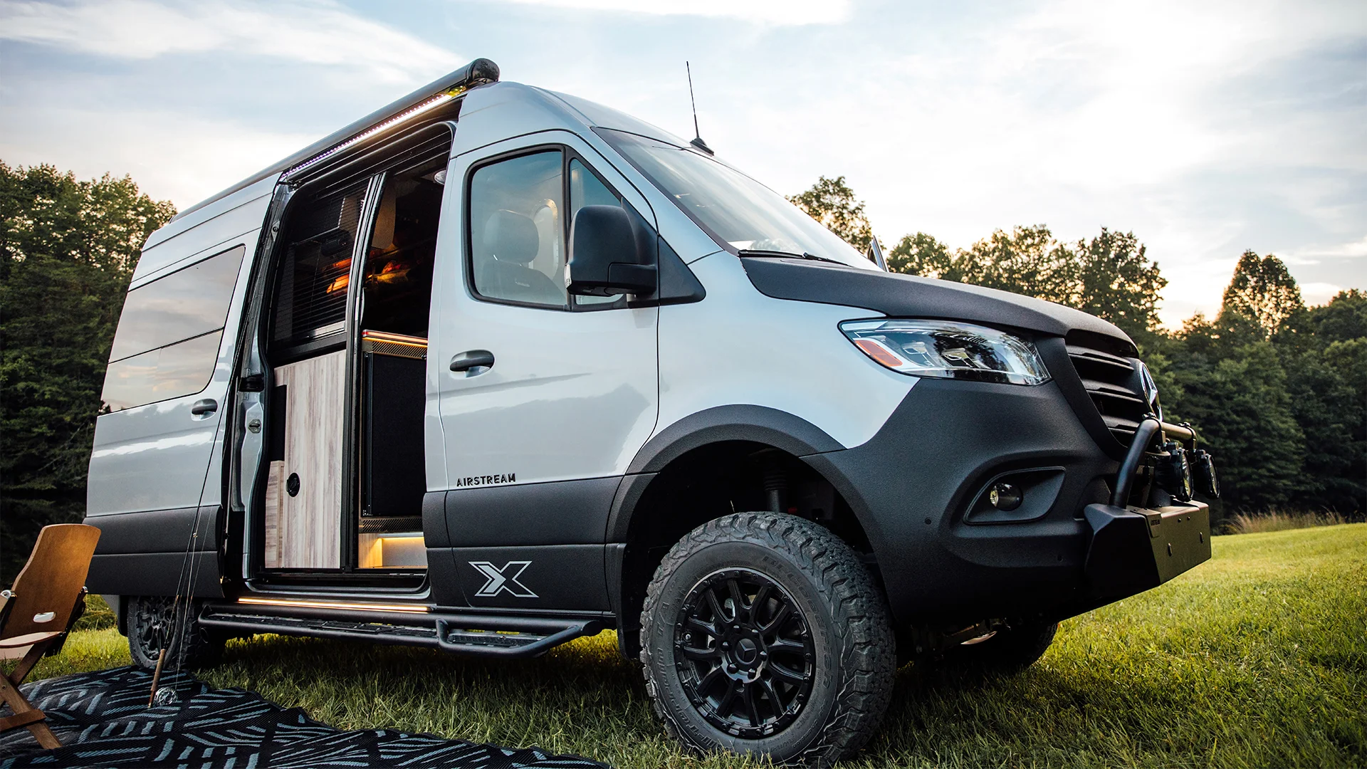 A rugged camper van with an open sliding door parked on grass, surrounded by trees under a blue sky with clouds.