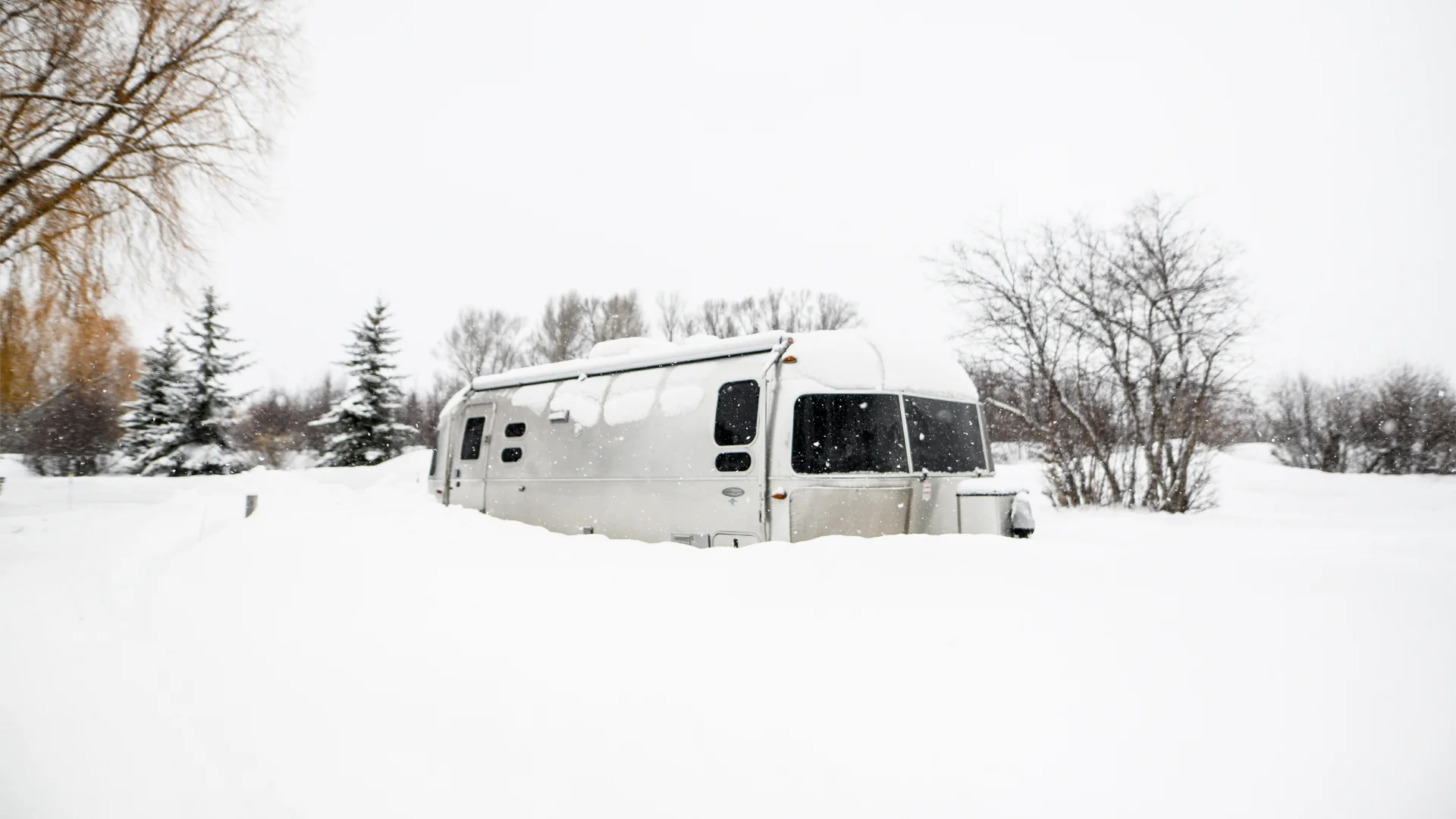 A silver trailer partially buried in deep snow, surrounded by leafless trees and pine trees, under a grey, overcast sky.