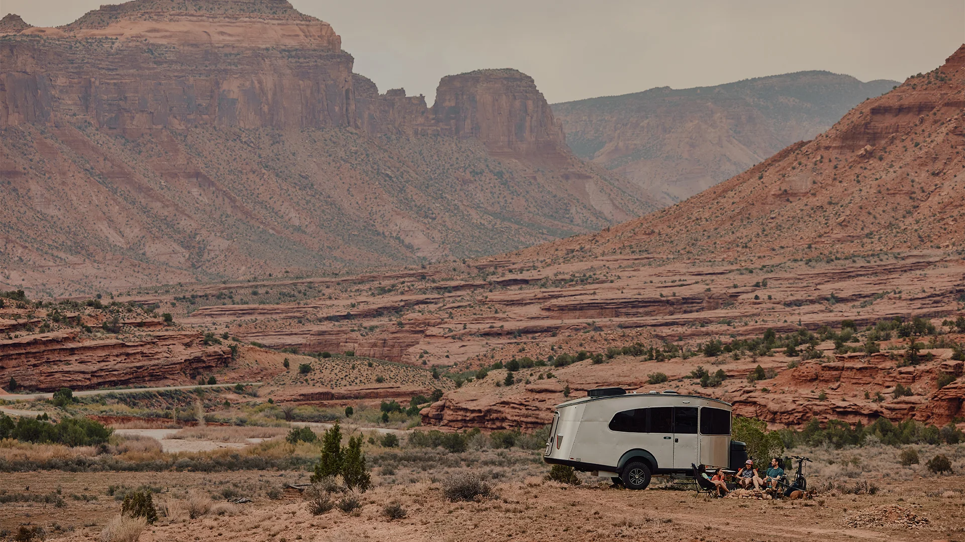 A camper trailer parked in a vast, rugged desert landscape with towering cliffs in the background, under a hazy sky.