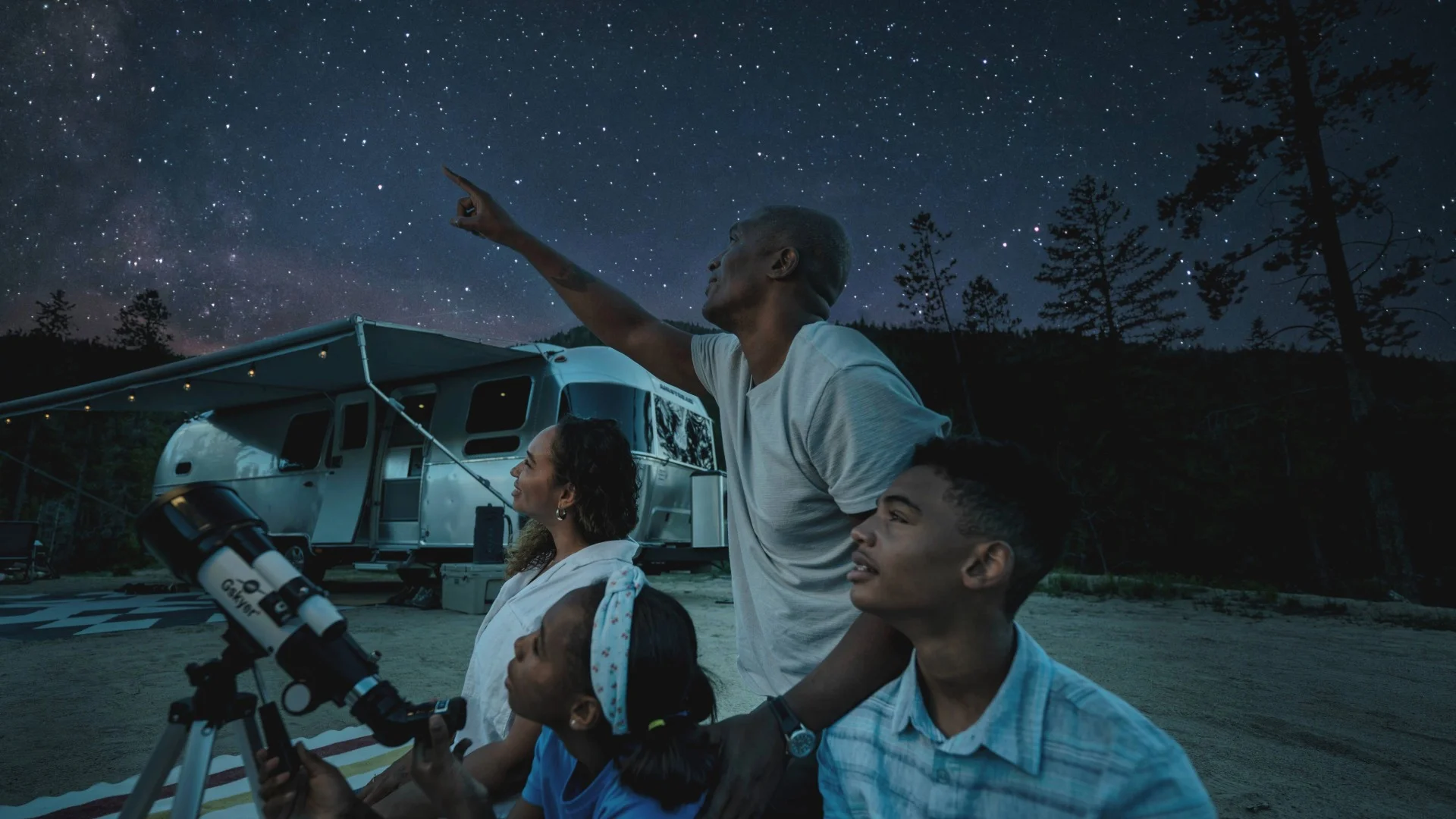 A family stargazing by an RV, one person pointing at the night sky filled with stars, while another looks through a telescope.