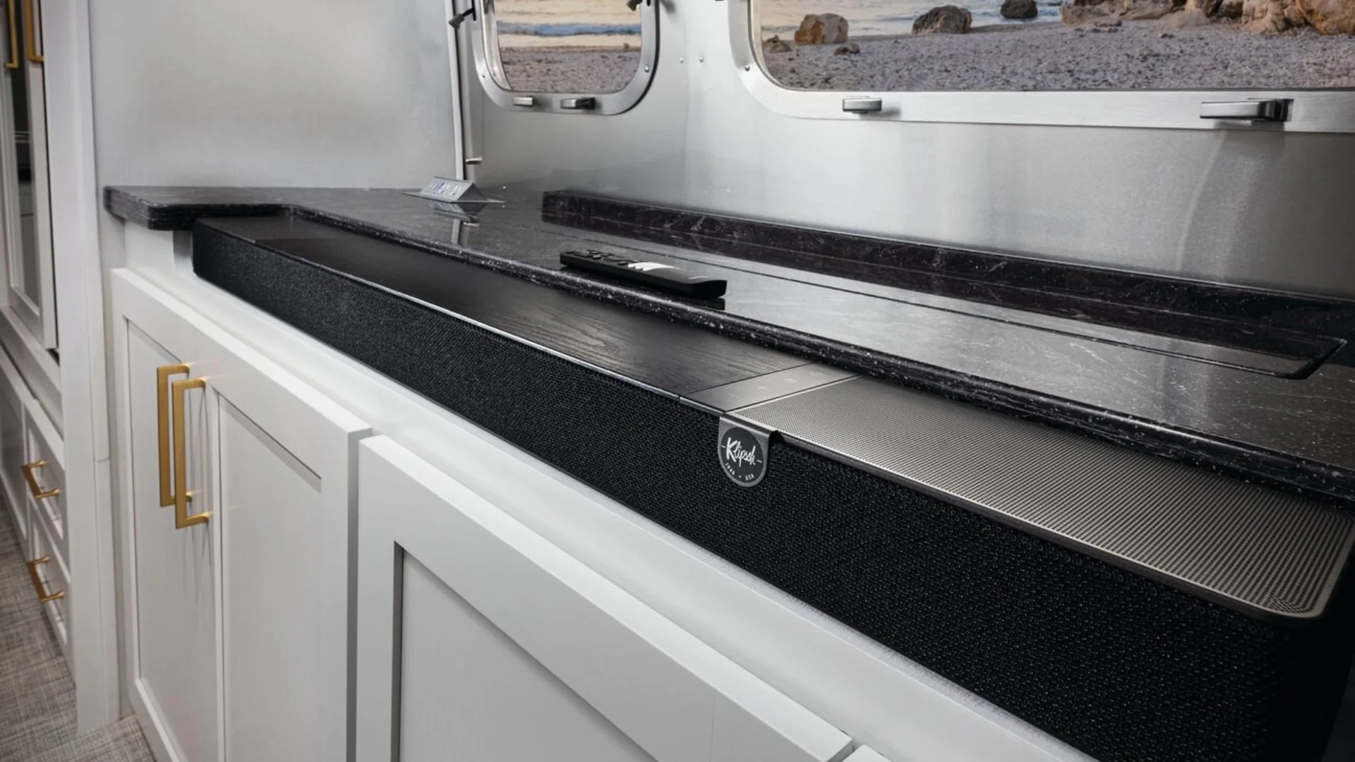 Modern kitchen counter in a motorhome with white cabinets, a sleek black stovetop, and gold handles, next to a window showing a beach view.