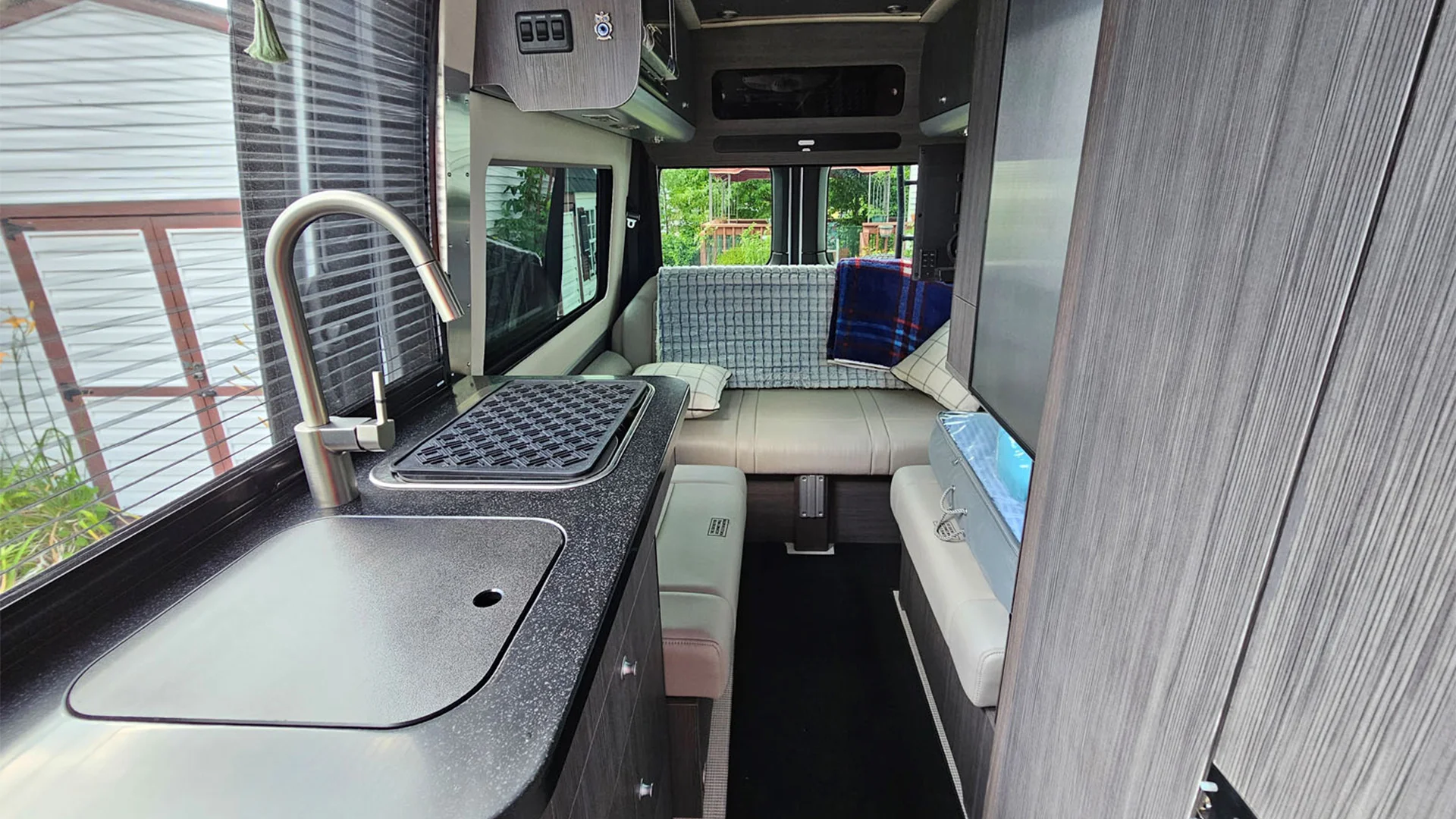 Interior of a modern camper van with a compact kitchen, seating area, and large windows. Light wood and dark countertops create a cozy atmosphere.
