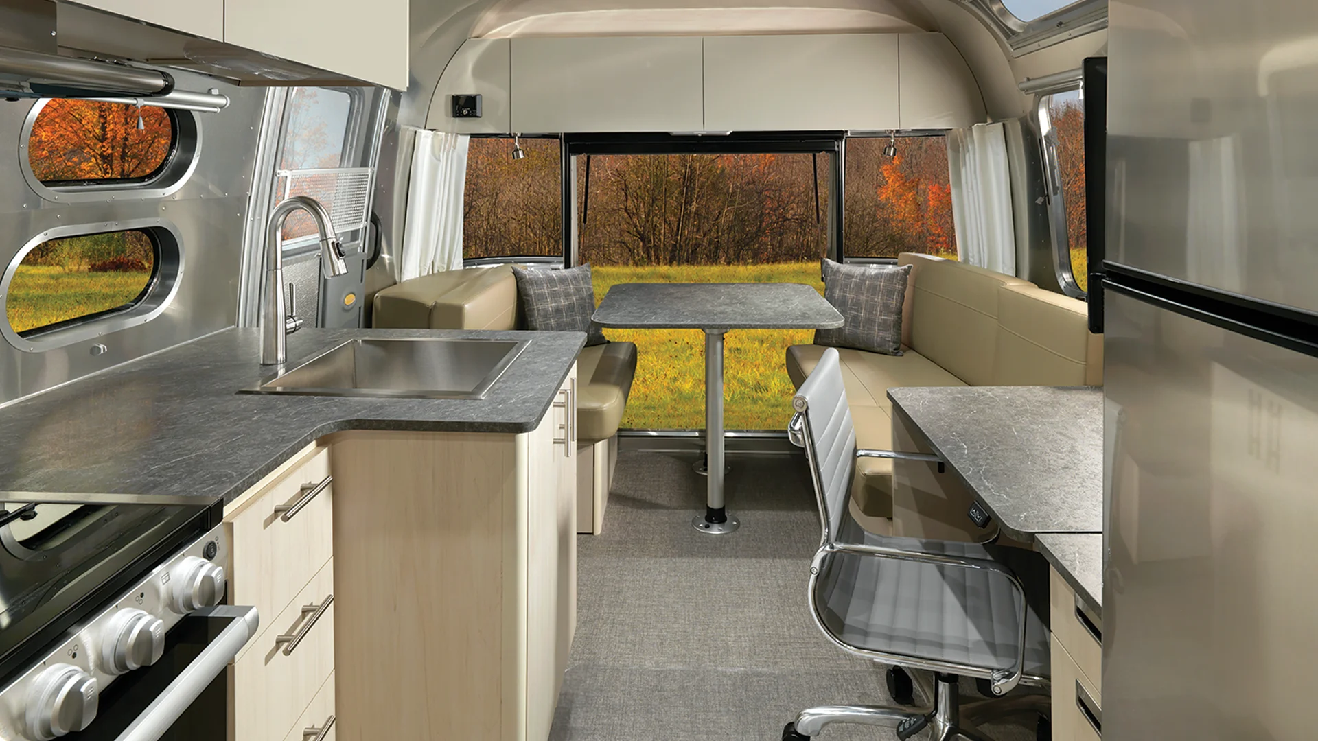 The interior of a modern RV features a kitchen with a stove, sink, and fridge, a dining area with a table and cushioned benches, an office chair at a work desk, and large windows showing a scenic autumn landscape.
