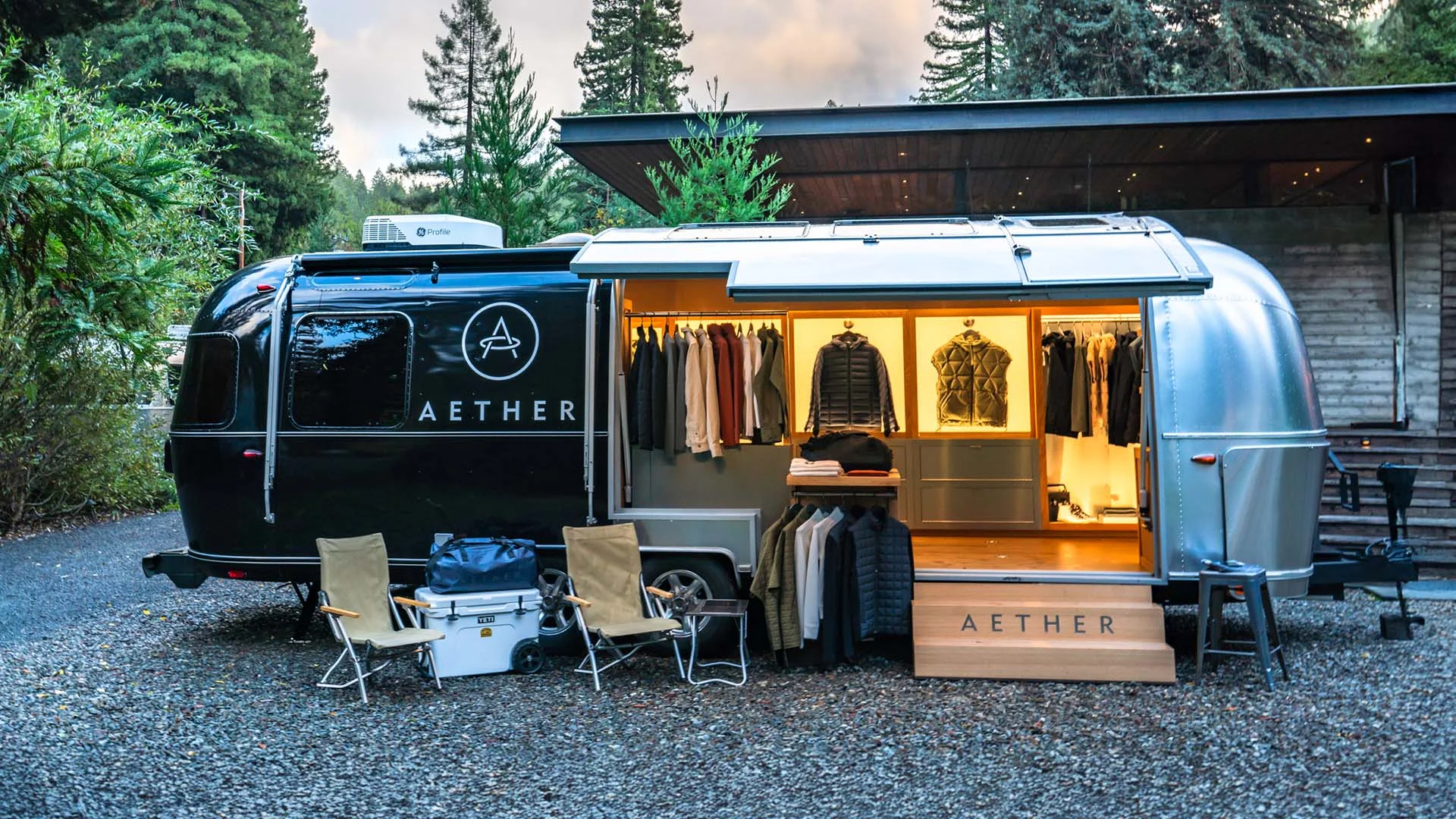 A sleek, silver Airstream trailer with "Aether" branding displays neatly arranged clothing, set in a wooded area with outdoor gear.