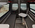 Interior of a camper van with two cushioned bench seats, a small foldable table between them, large windows on all sides, and a view of a sandy, rocky landscape outside.