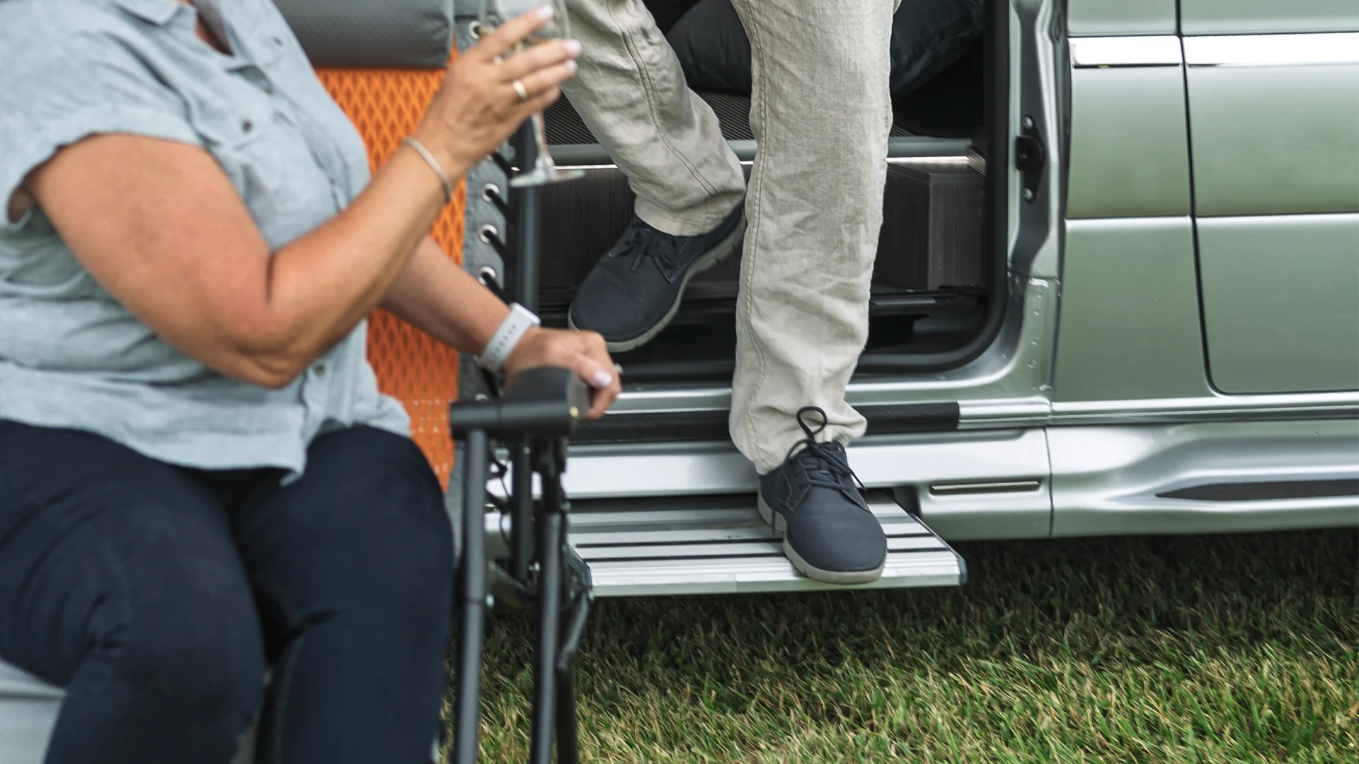 A person steps out of a vehicle onto a retractable step, while another person sits nearby on a foldable chair on grass.