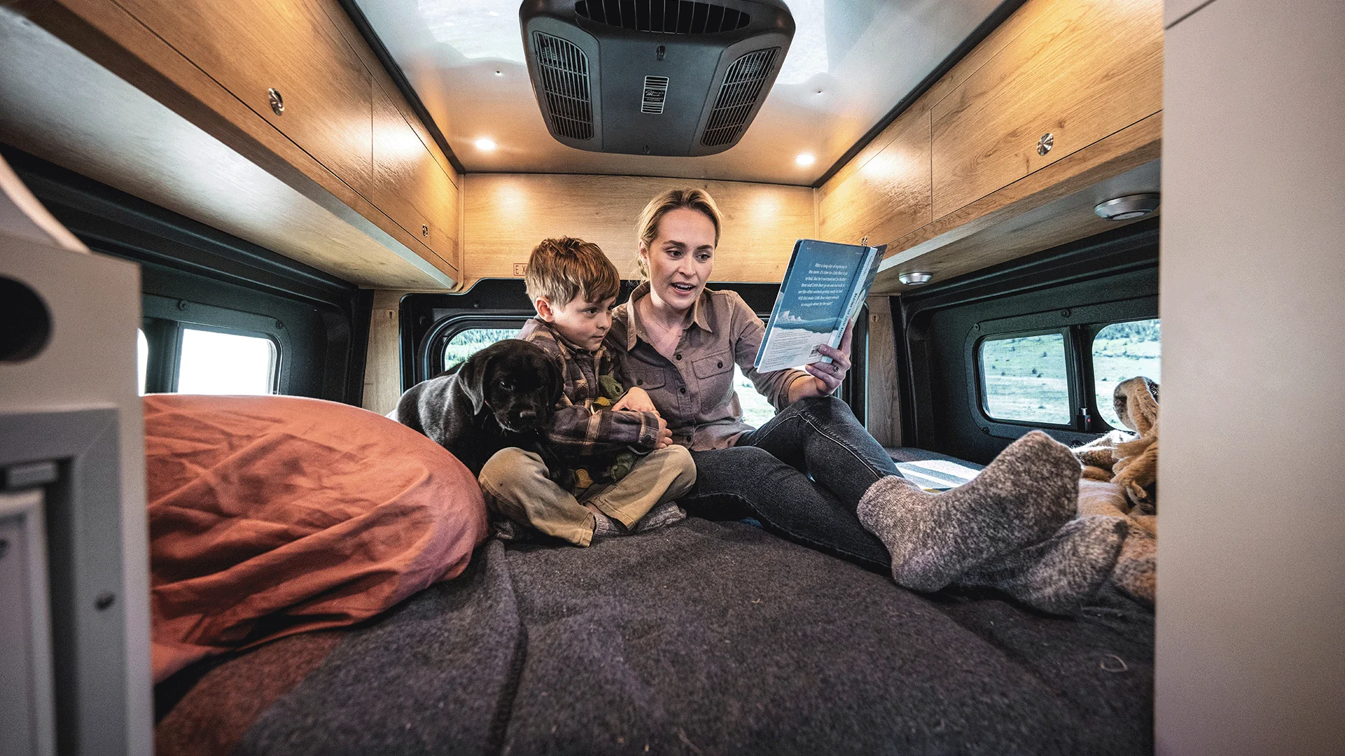 A woman reads a book to a child and a dog inside a cozy camper van, with wood paneling and soft lighting, creating a warm, inviting atmosphere.