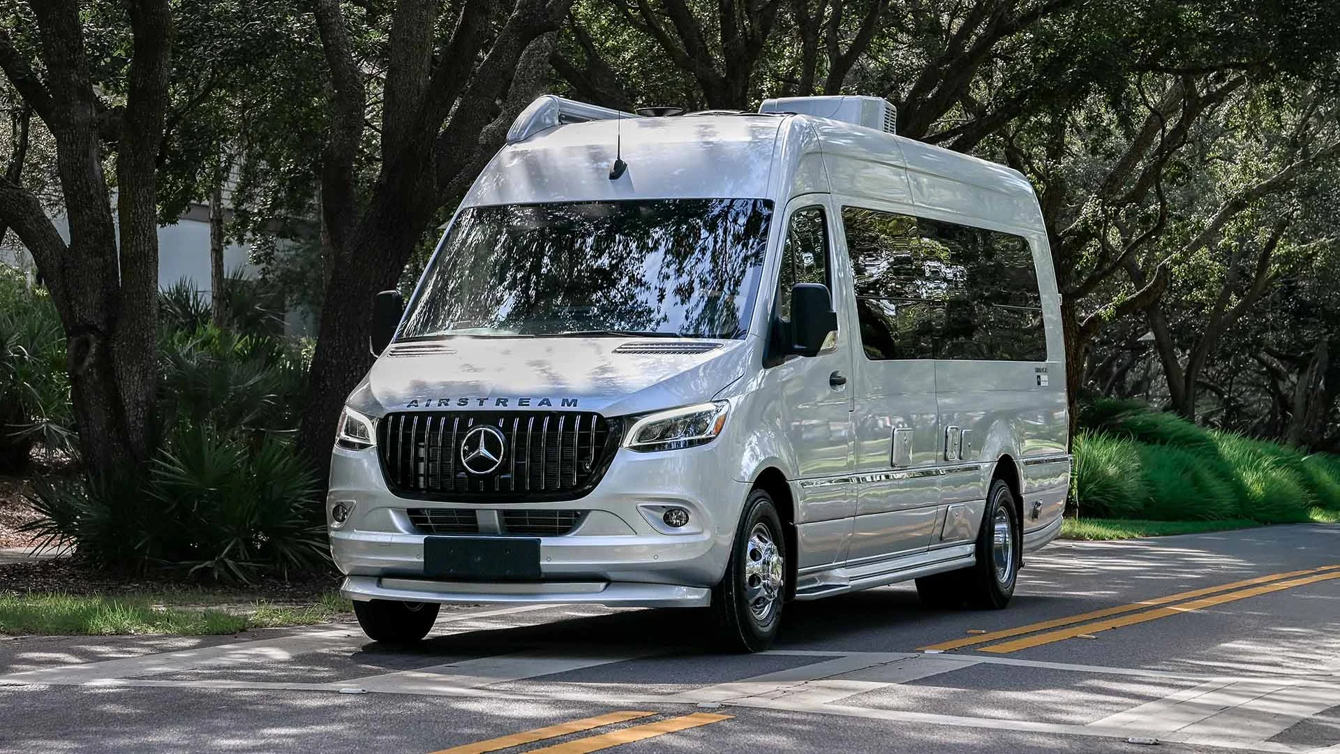 White Mercedes-Benz Sprinter van on a tree-lined road, featuring a sleek design and tall body, suitable for travel or transport.