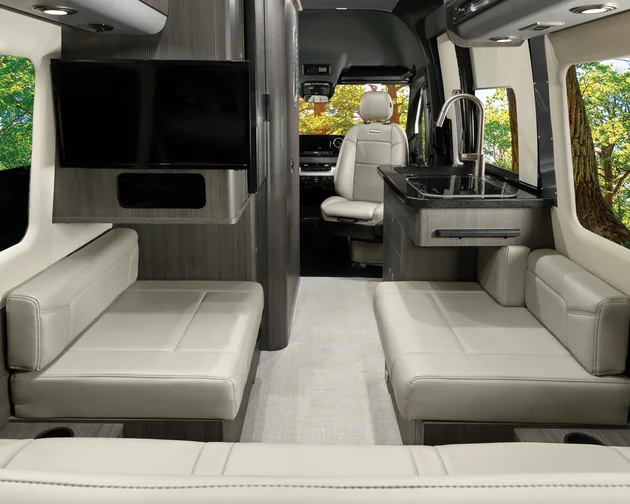 Interior of a modern camper van featuring cream leather seating, a TV, a small kitchenette with a sink, wood paneling, and large windows showing a forested outdoor scene.
