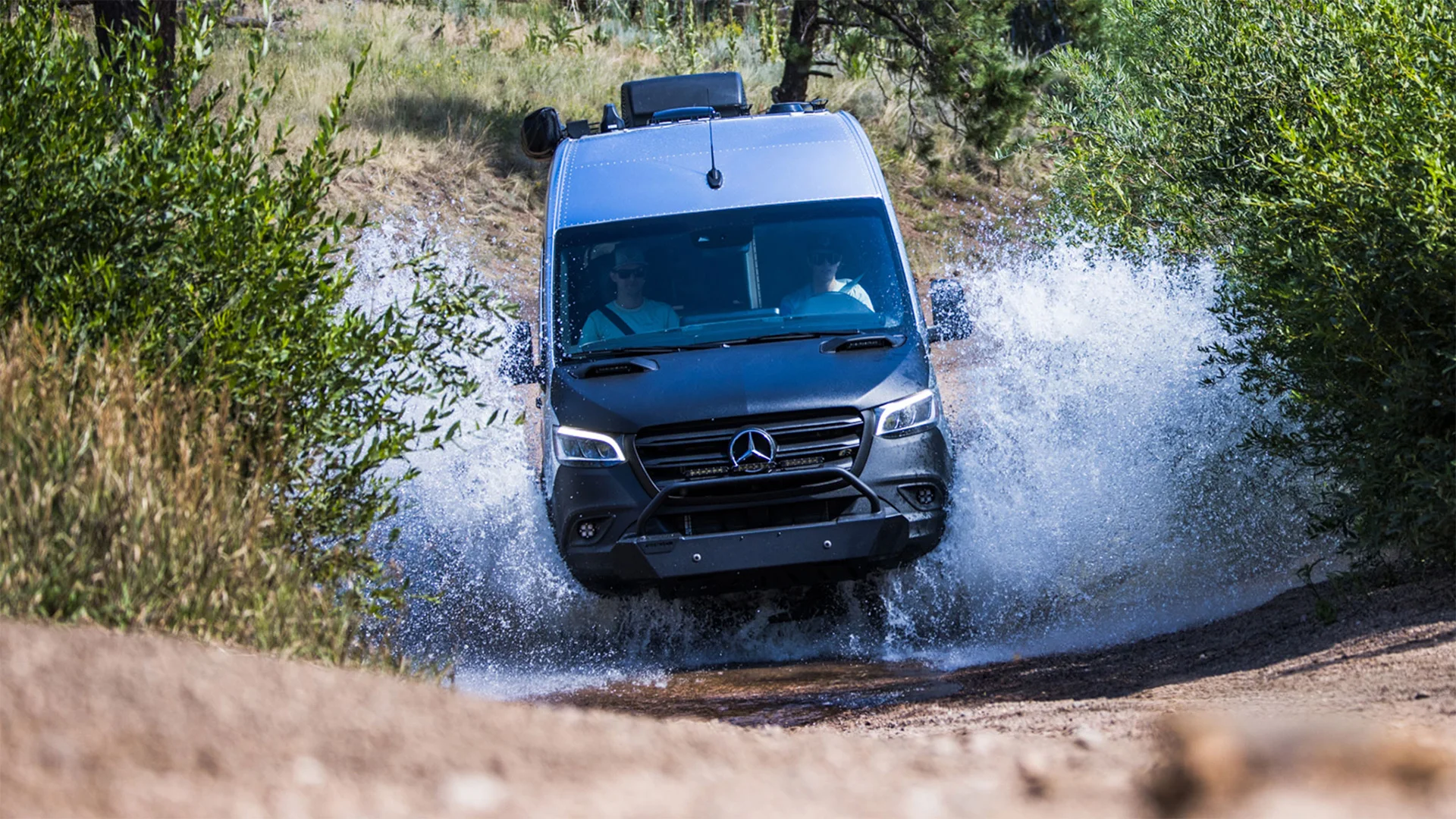 A Mercedes-Benz van drives through a water splash on a dirt path, surrounded by greenery and trees in a rugged outdoor setting.