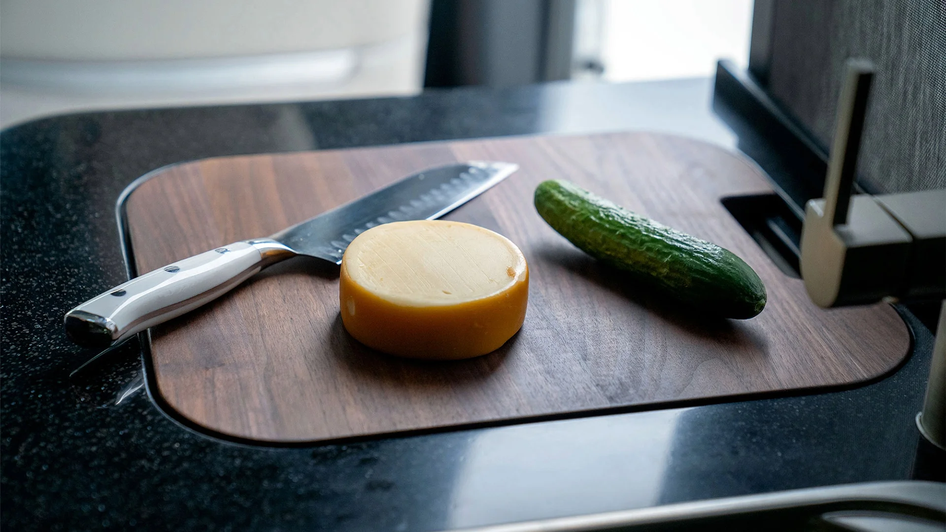 Cheese wheel, cucumber, and knife on a wooden cutting board atop a dark countertop in a kitchen setting.