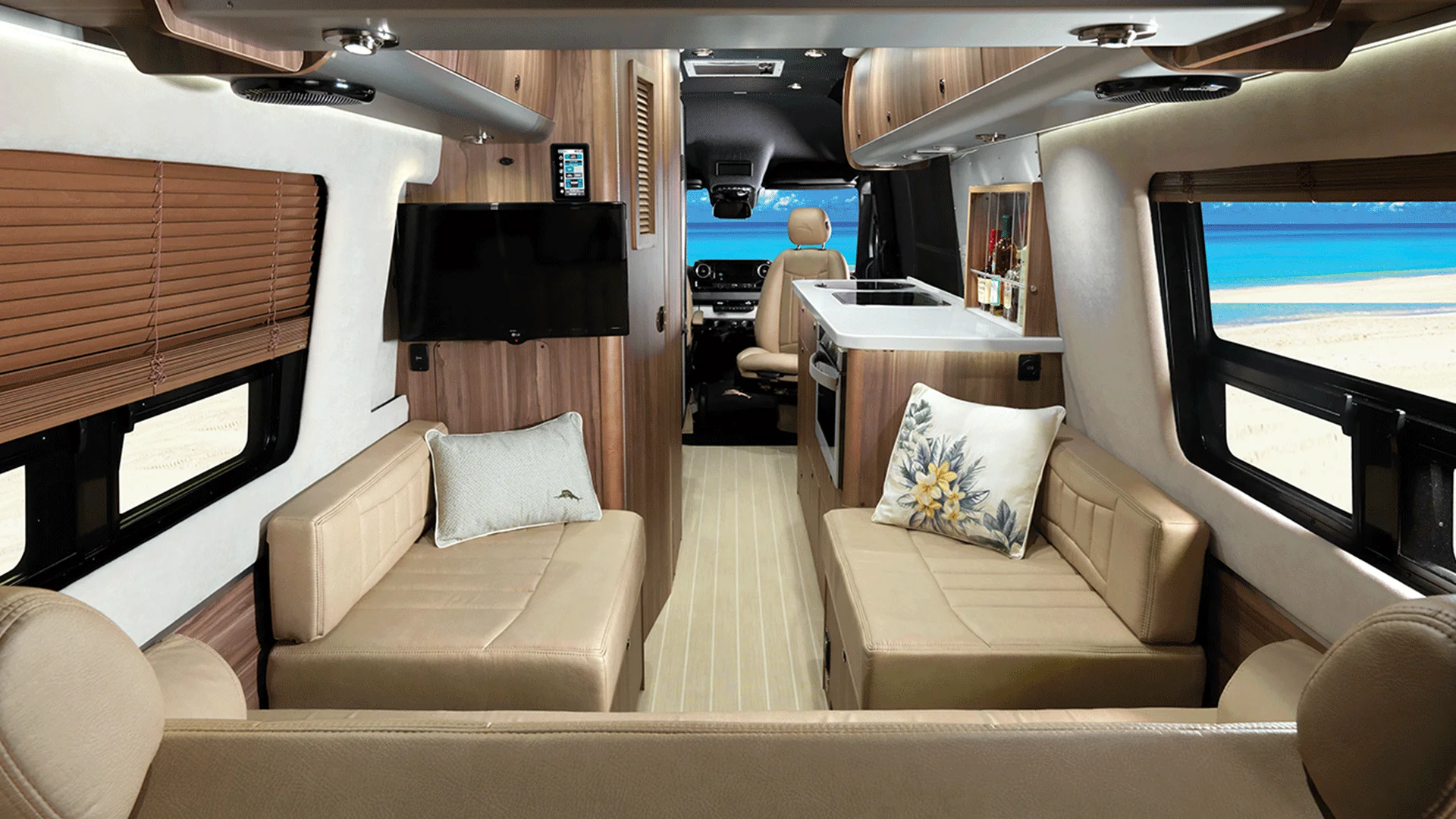 Luxury camper van interior with beige leather seating, a small kitchen, and windows overlooking a beach with blue water.