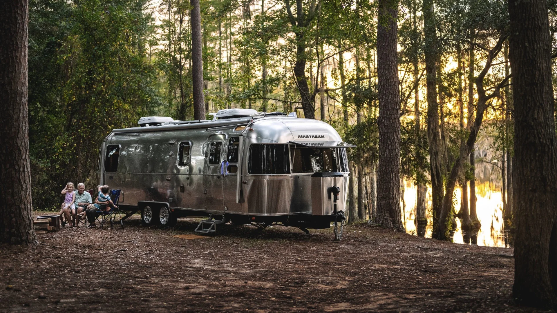 A shiny Airstream trailer parked in a forested campsite near a lake, with people sitting outside enjoying the evening among tall trees.