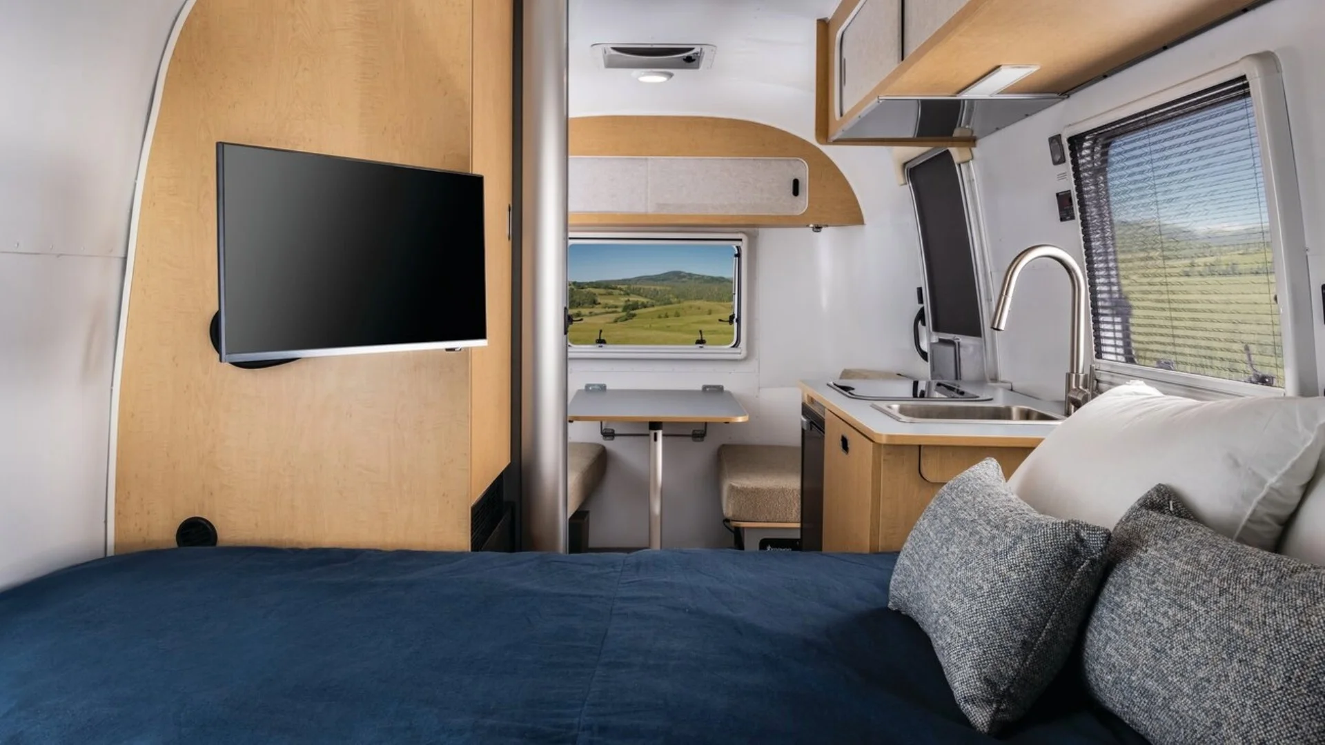 Interior of a modern campervan with a cozy bed, small kitchen, and dining area. Large windows reveal a scenic view of rolling hills outside.