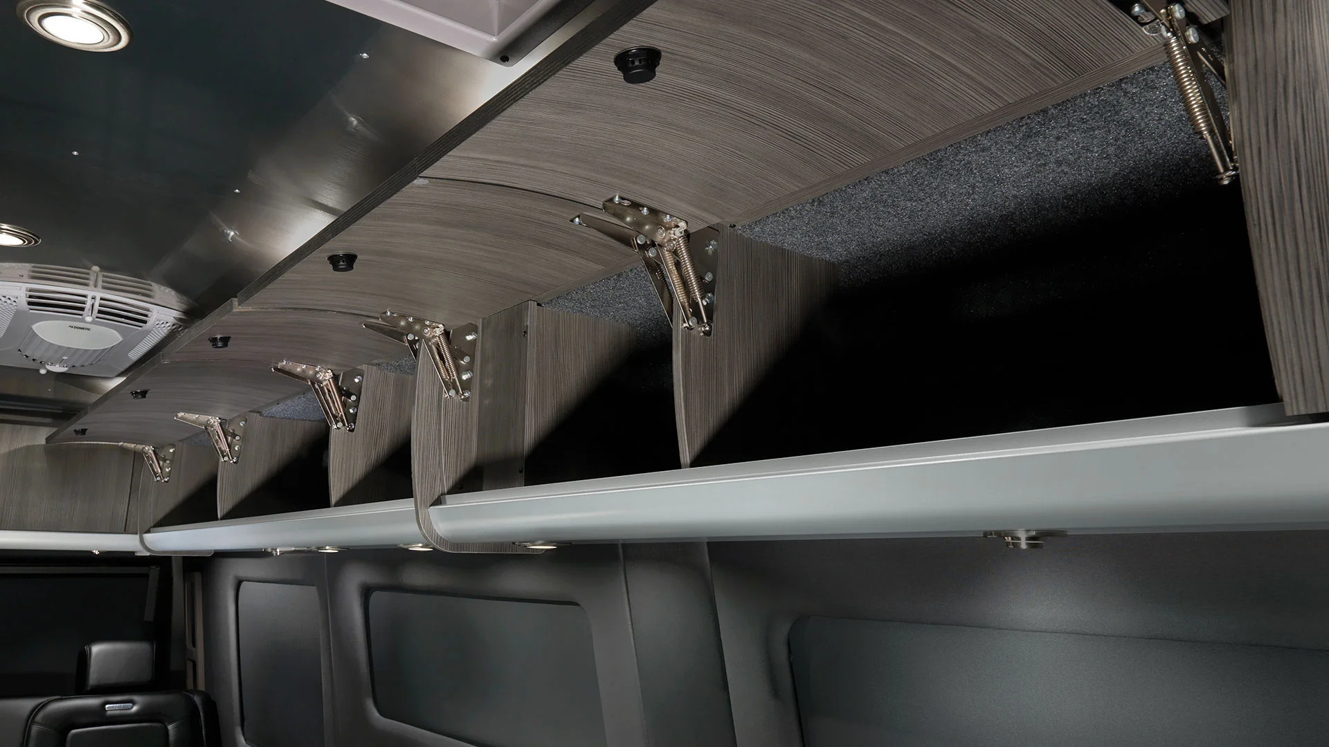 Interior view of a vehicle showcasing open overhead storage compartments with wood paneling and metal hinges, featuring dark upholstered seats below.