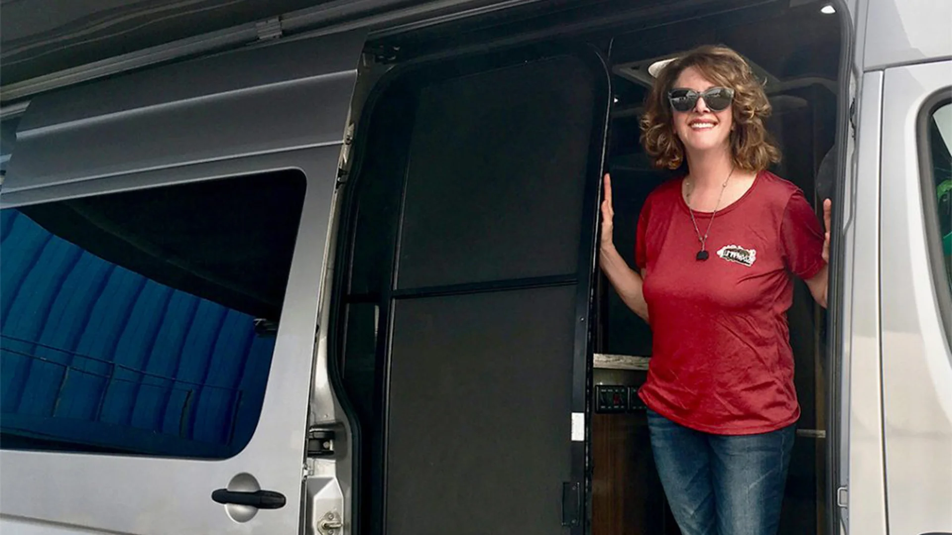 Woman in sunglasses and a red t-shirt stands at the open door of a silver van, smiling cheerfully.