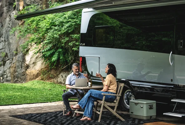 A couple sits outside an RV with a laptop and drinks, surrounded by lush greenery and rocky terrain.