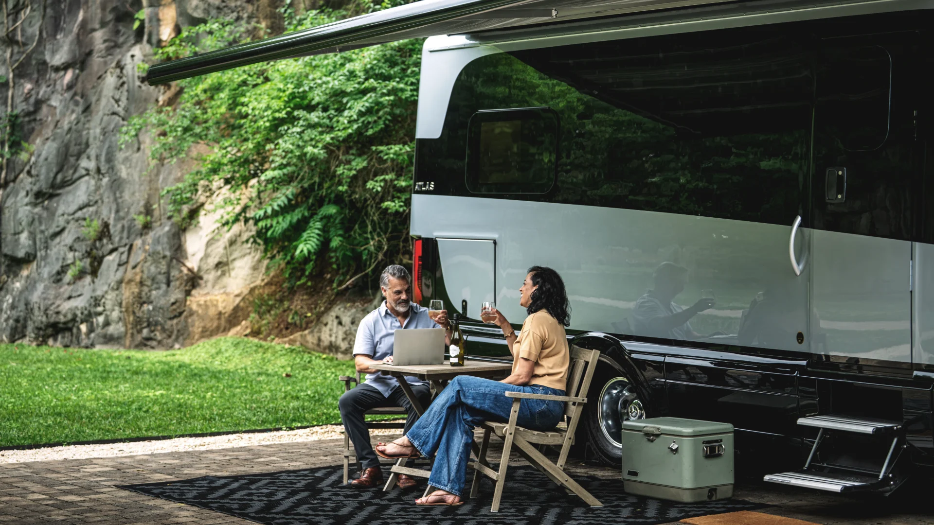 A couple sits outside an RV with a laptop and drinks, surrounded by lush greenery and rocky terrain.
