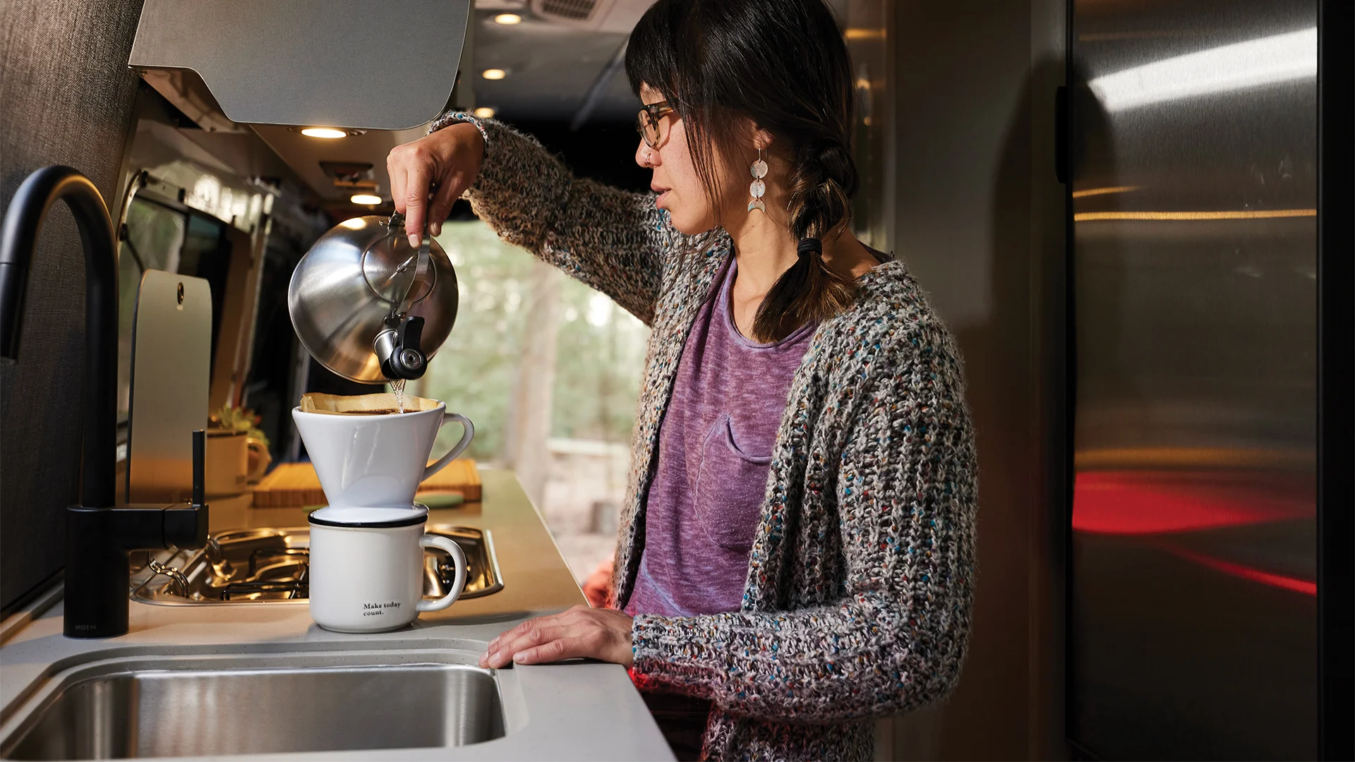Woman pouring water from a kettle into a coffee dripper in a modern kitchen with a stainless steel fridge and dark cabinetry.