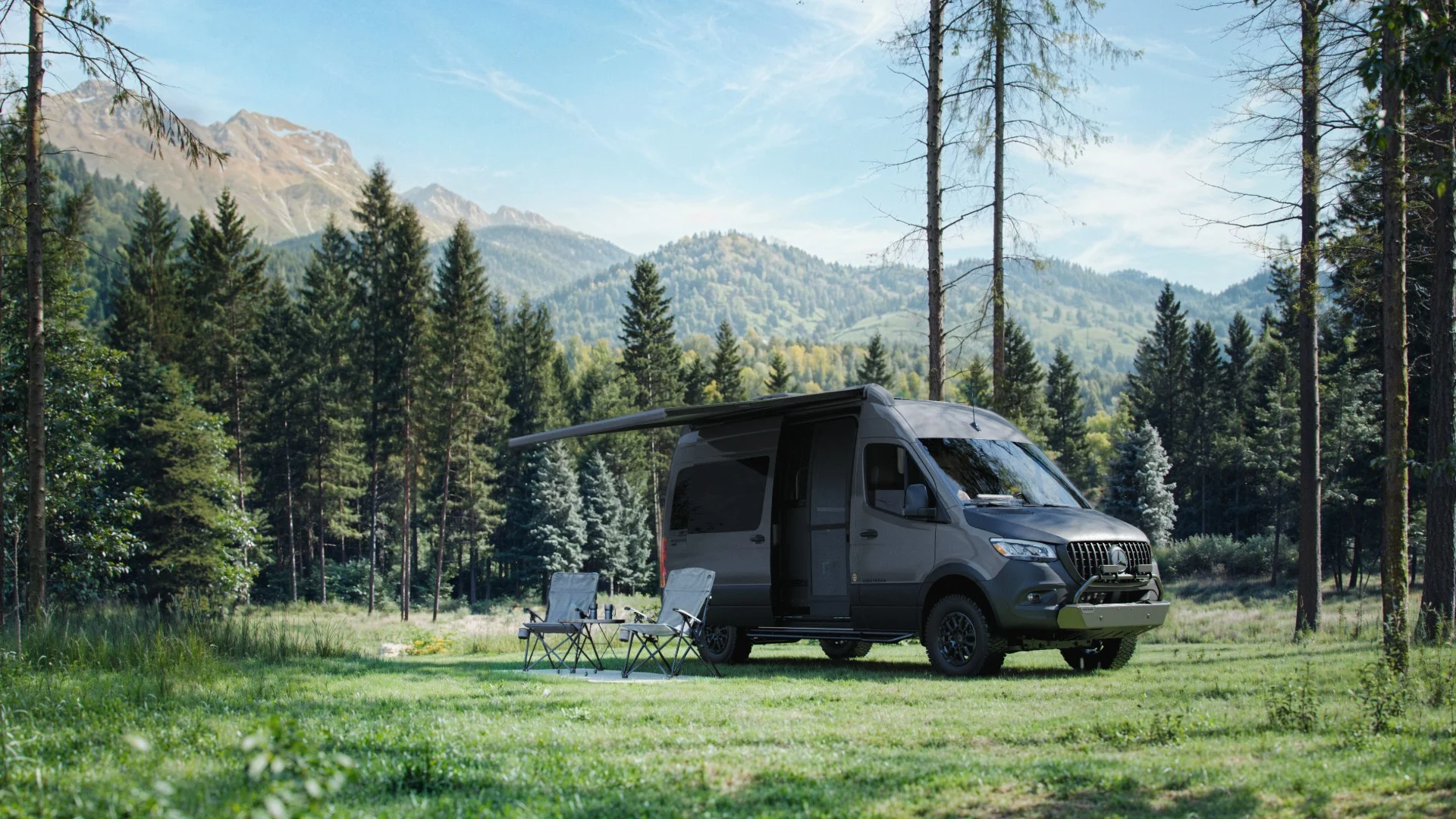 A camper van with an extended awning is parked in a grassy clearing surrounded by tall trees, with mountains in the background. Camping chairs are set up nearby.