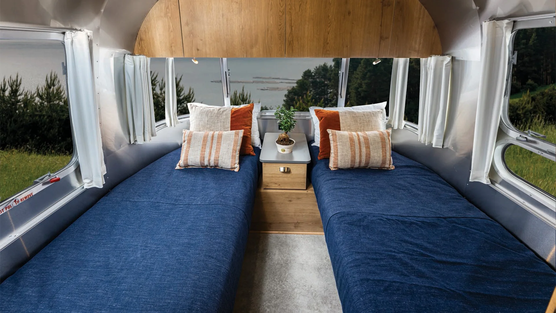 Interior of a camper with two blue twin beds, white and orange pillows, a wooden nightstand with a potted plant, and a window view of nature.