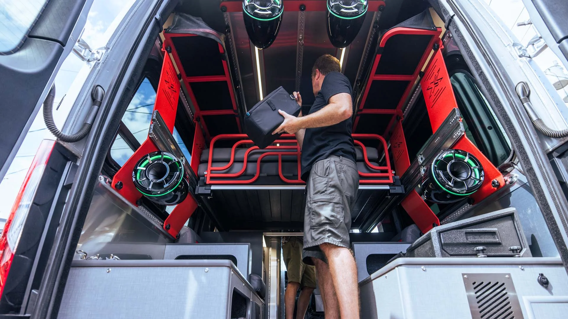 Man organizing equipment inside a van converted into a mobile workshop, with red shelves and green speakers.