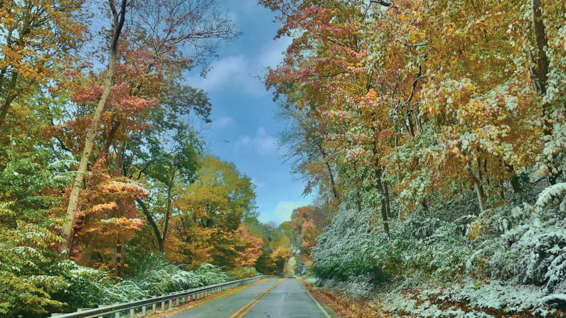 A scenic road lined with trees showing autumn foliage in shades of orange, yellow, and red, with some branches and ground covered in a light layer of snow under a partly cloudy sky.