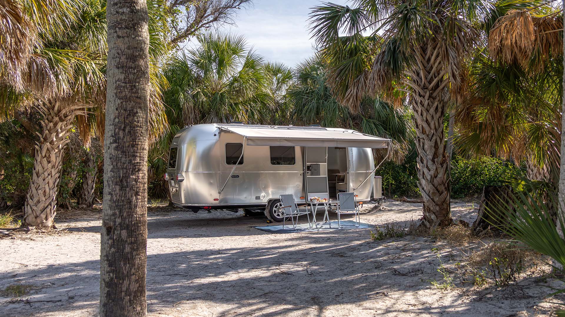 A silver RV is parked among palm trees on a sandy ground, with two folding chairs and a small table set up outside.