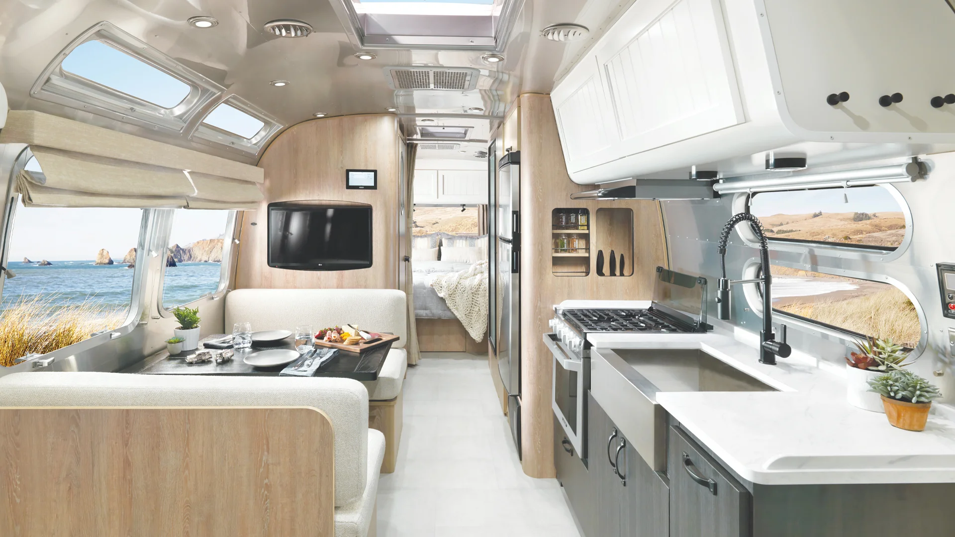 Modern RV interior with light wood and white decor, featuring a dining area set with food, kitchen with stove and sink, and a bed in the background; large windows show a scenic coastal view.