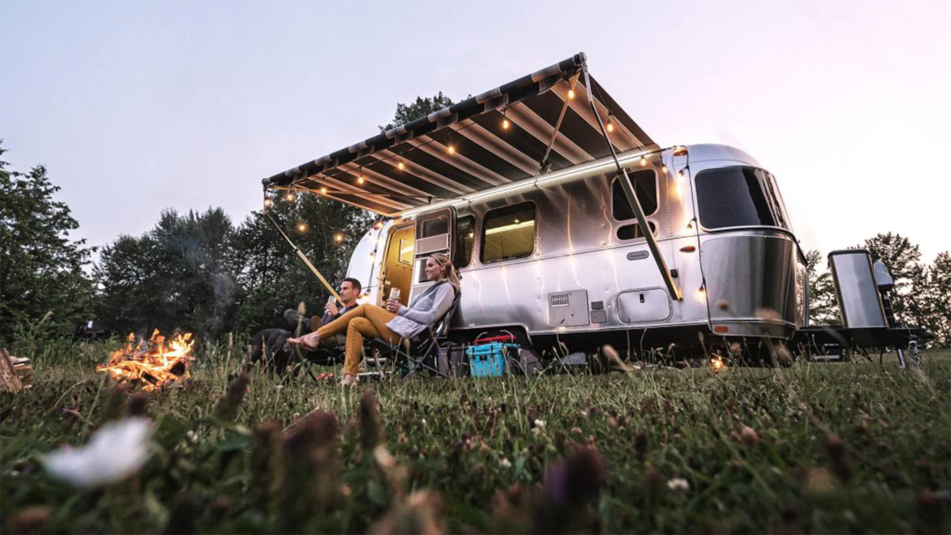Couple relaxes outside a shiny trailer with an awning and string lights, near a campfire, in a grassy area at dusk.