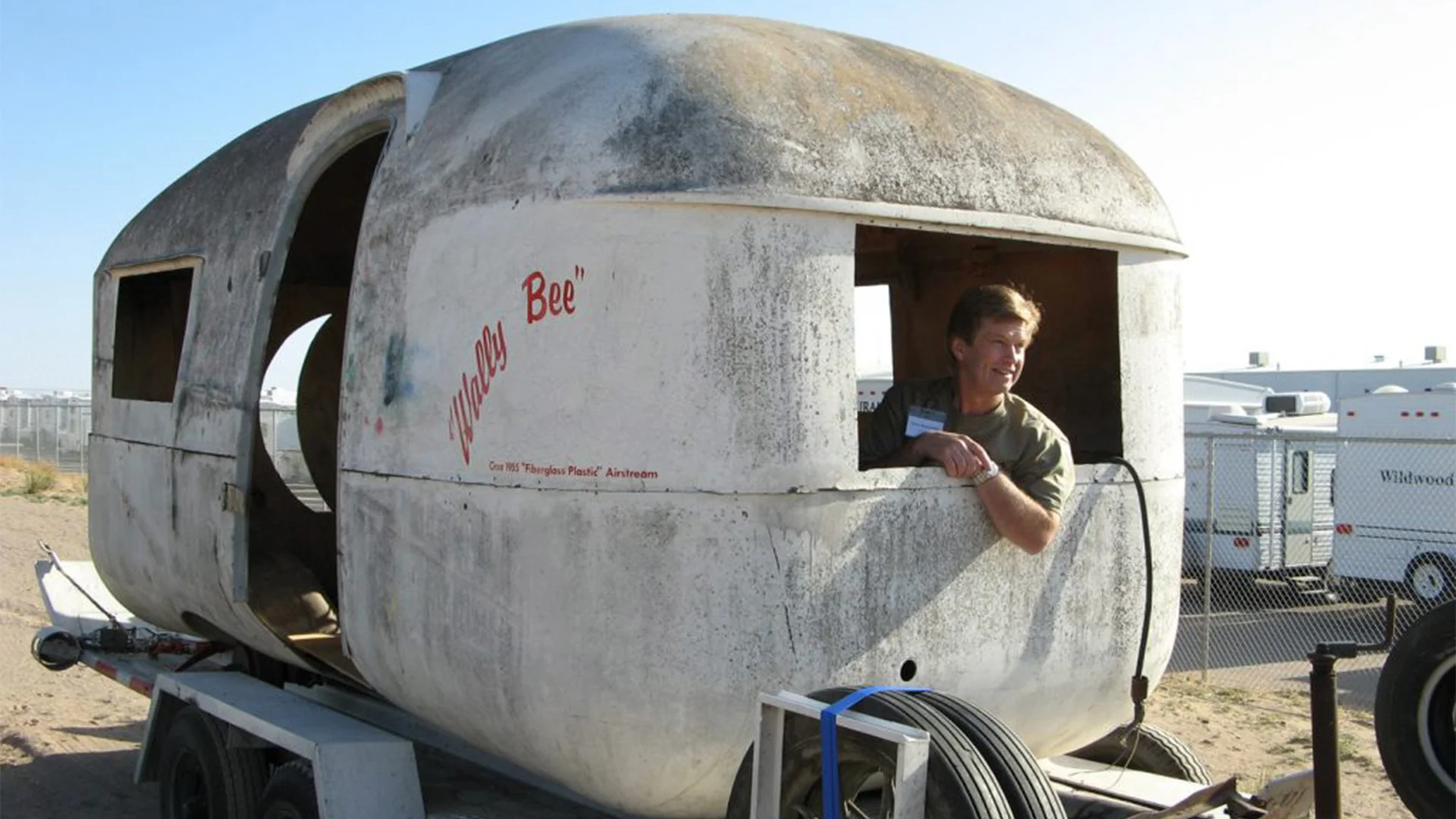Man leaning out of a weathered, oval-shaped camper on a trailer, with "Wee-ite Bee" text, in a parking area with other trailers.