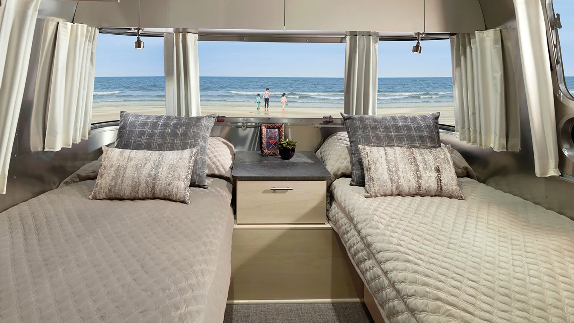 Interior of an Airstream trailer with twin beds, open curtains, and a view of a beach and ocean through a large window.