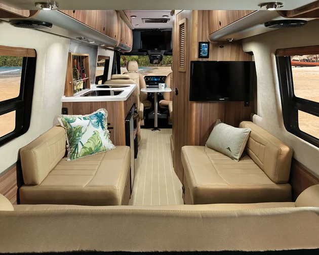 Interior view of a modern RV with tan leather seating, decorative pillows, a small kitchen area, mounted TV, large windows, and a beach visible outside. The space looks bright, organized, and inviting.