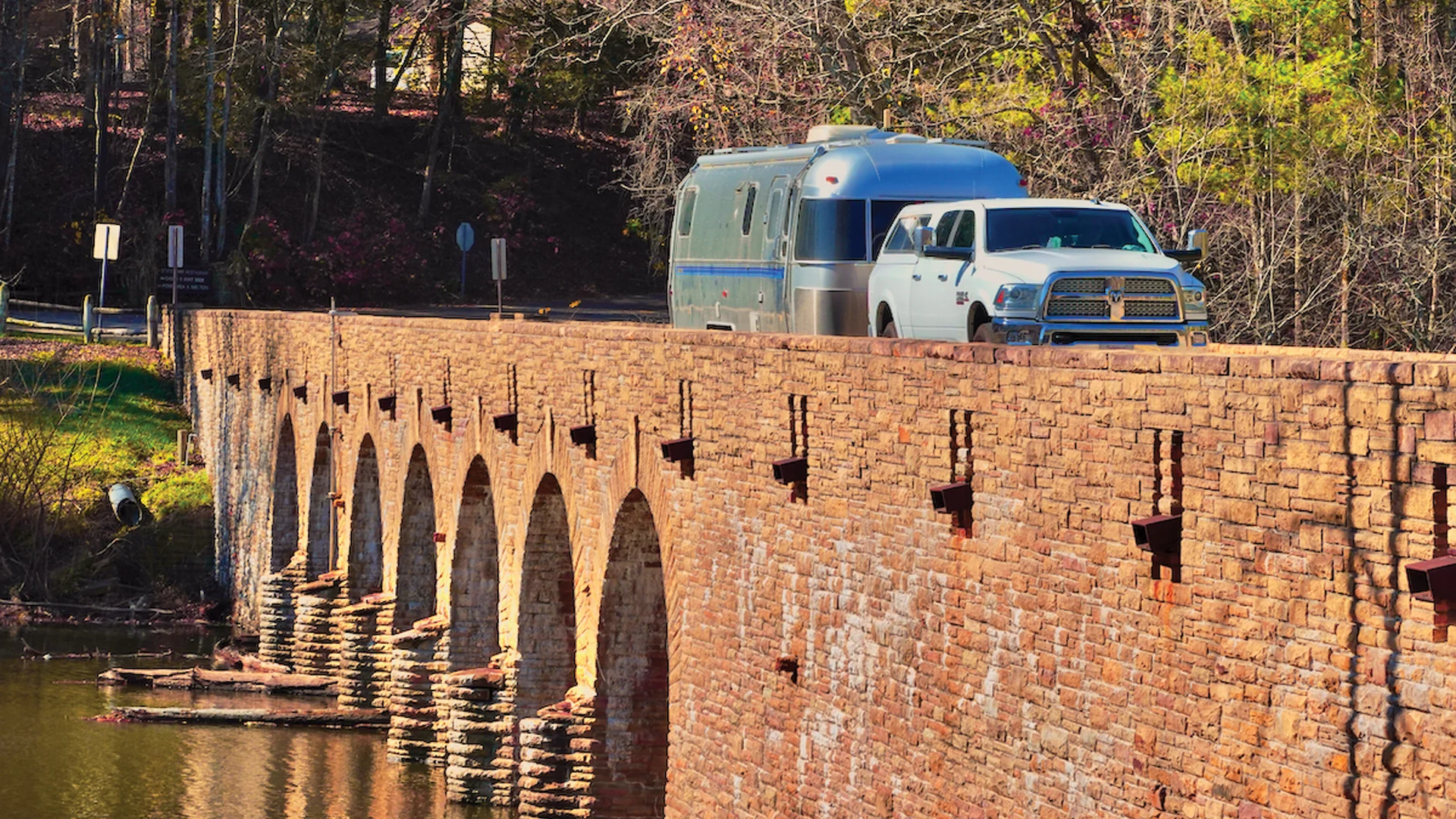 A white pickup truck towing a silver camper trailer crosses a historic stone arched bridge over a calm river, surrounded by trees with early spring foliage.