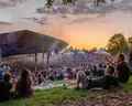 Crowd at Bethel Woods Performing Arts Center, seated on the grass, enjoying music under a colorful sunset sky.