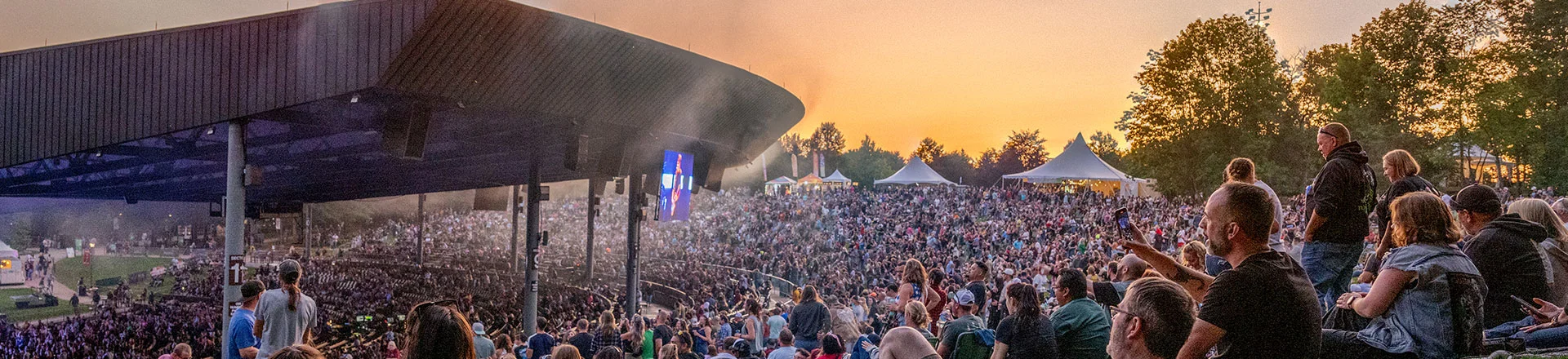 Crowd at Bethel Woods Performing Arts Center, seated on the grass, enjoying music under a colorful sunset sky.