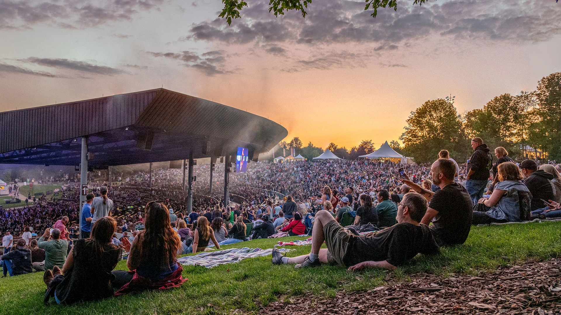 Crowd at Bethel Woods Performing Arts Center, seated on the grass, enjoying music under a colorful sunset sky.