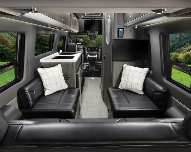 Interior of a luxury camper van with black leather seating, plaid pillows, a small kitchen area with a sink, flat-screen TV, large windows showing green countryside and trees, and the drivers cab visible at the front.
