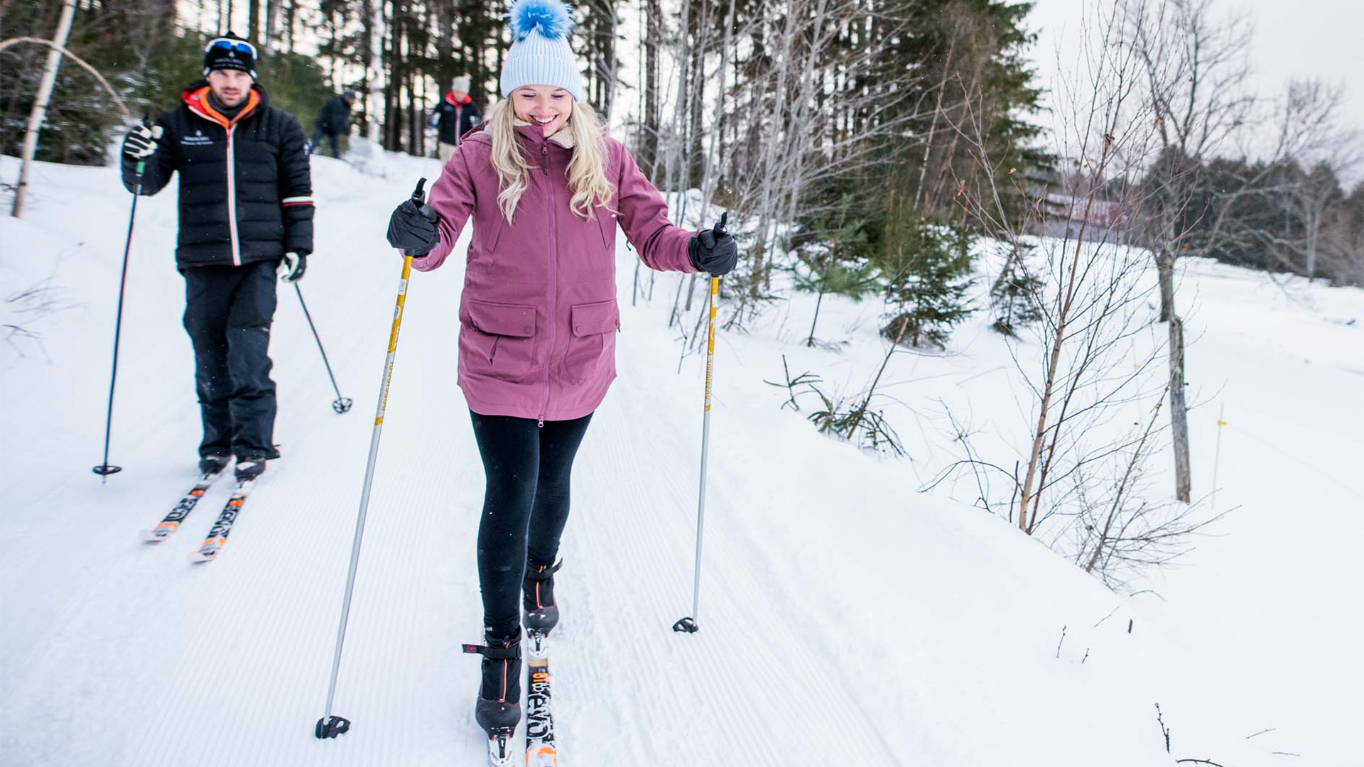Two people cross-country skiing on a snowy trail, surrounded by trees. The person in front wears a purple coat and a blue pom-pom hat.