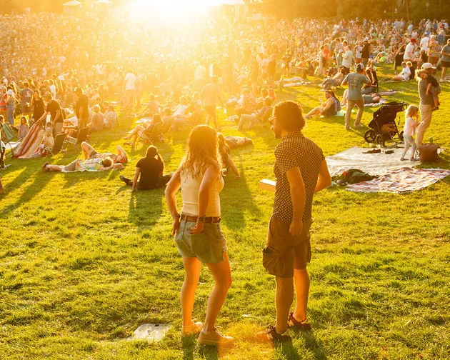 Crowd enjoying an outdoor event on a sunny day, with people sitting on blankets and grass, and a golden sunset illuminating the scene.