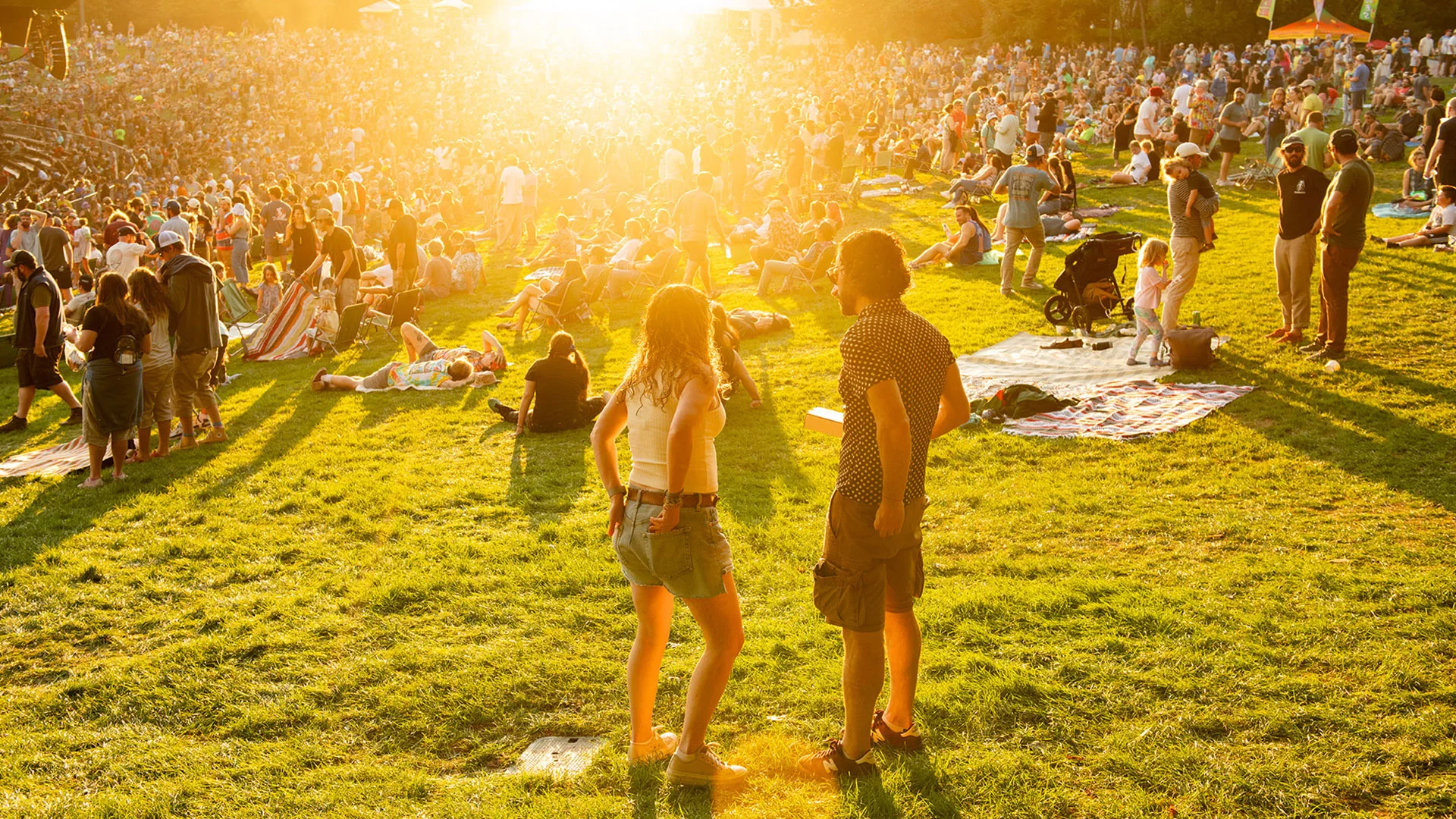 Crowd enjoying an outdoor event on a sunny day, with people sitting on blankets and grass, and a golden sunset illuminating the scene.