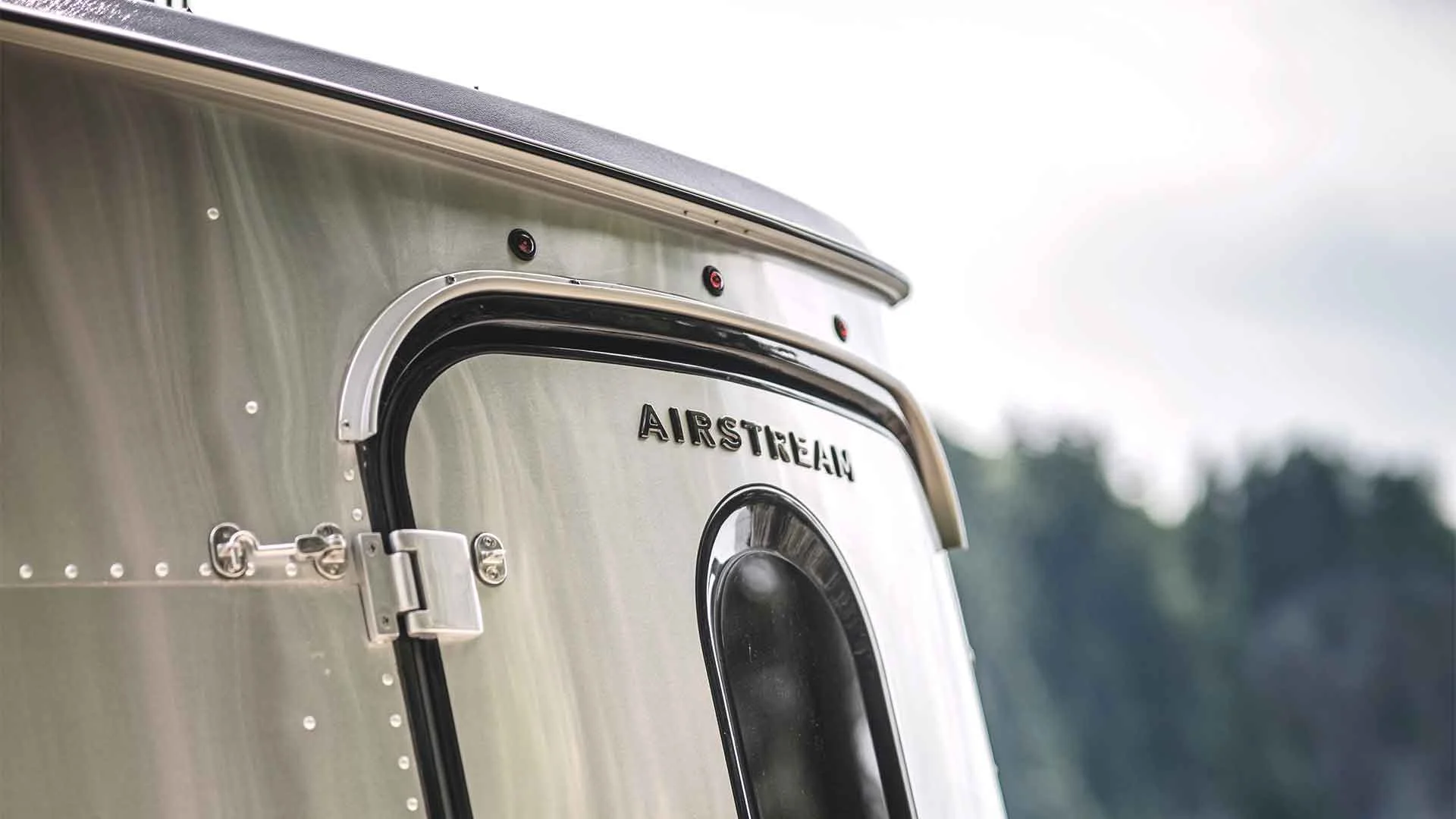 Close-up of an Airstream trailer corner showing its polished aluminum exterior and door with a blurred forest in the background.