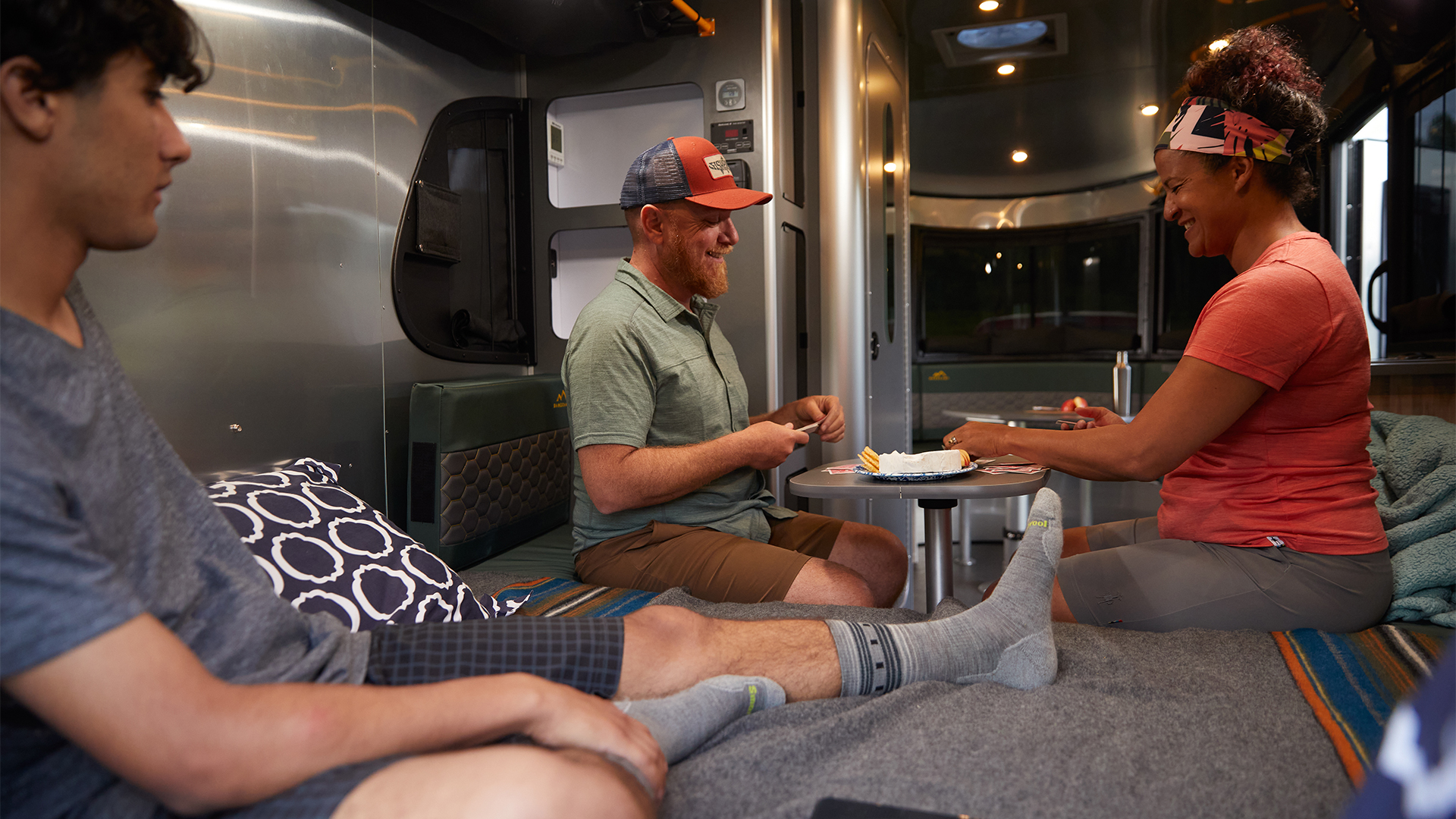 Three people relaxing inside a trailer, sitting around a small table with snacks, one person reclining with legs outstretched on a bed.