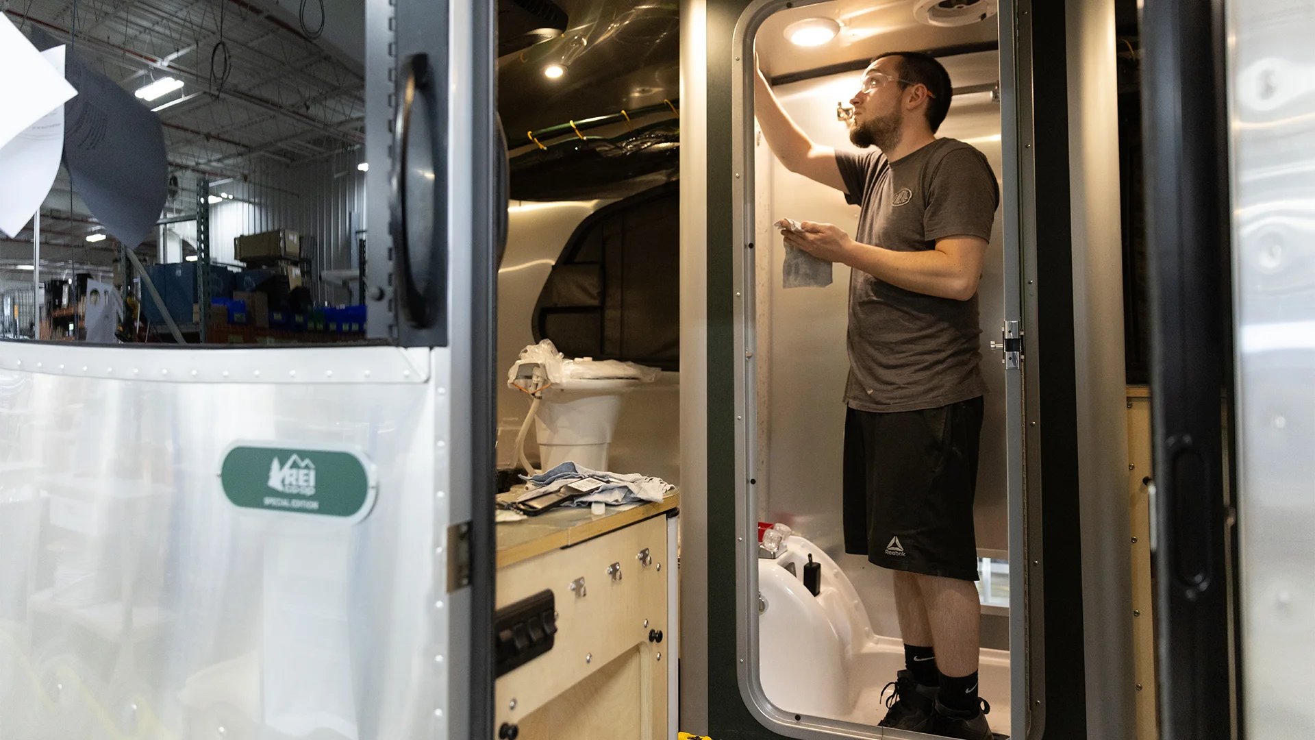 Man inspecting the interior of an RV bathroom, checking a vent or light, in a workshop setting with various tools visible.