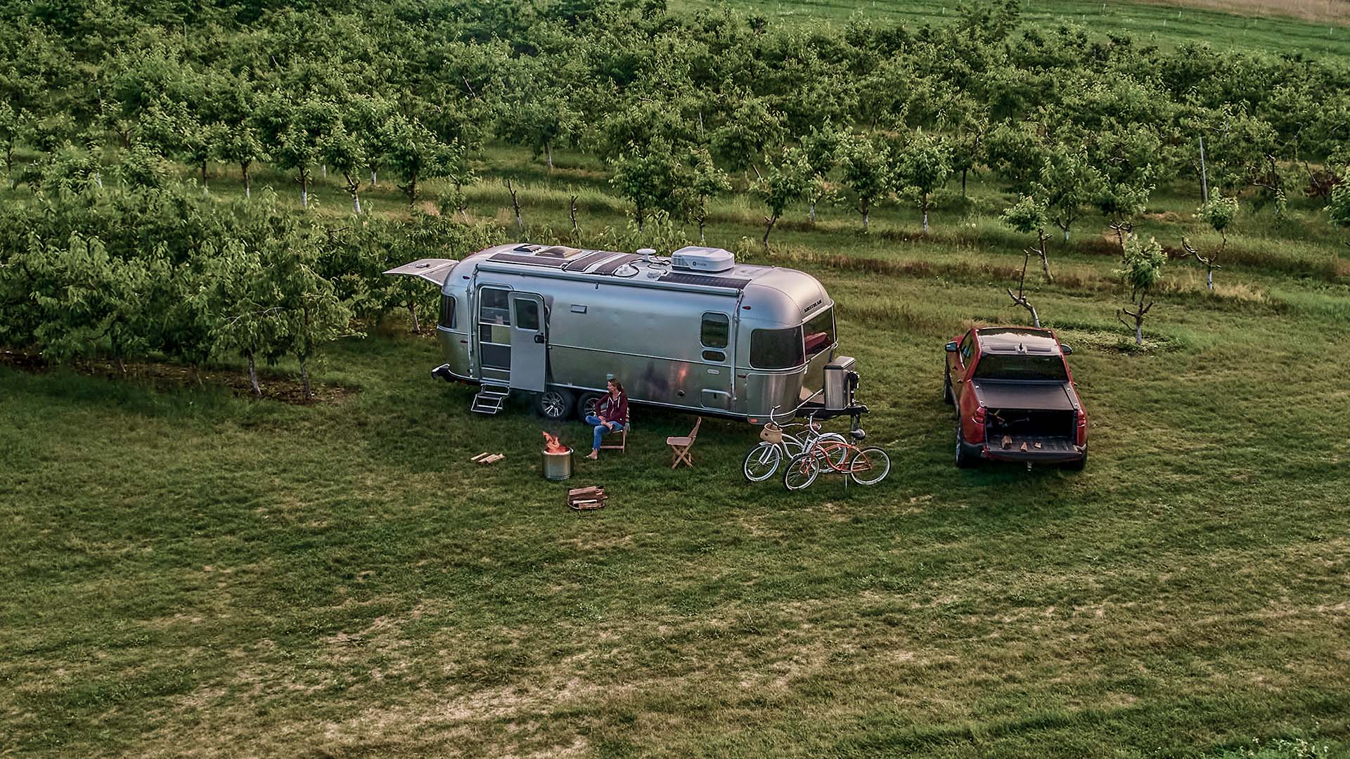 Aerial view of an Airstream trailer and pickup truck parked on grass near rows of trees, with bicycles, chairs, and a campfire setup outside.