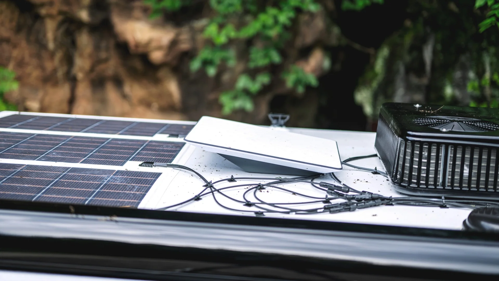 A solar panel system and a satellite dish are mounted on the roof of a vehicle, with cables and a fan unit, surrounded by greenery.