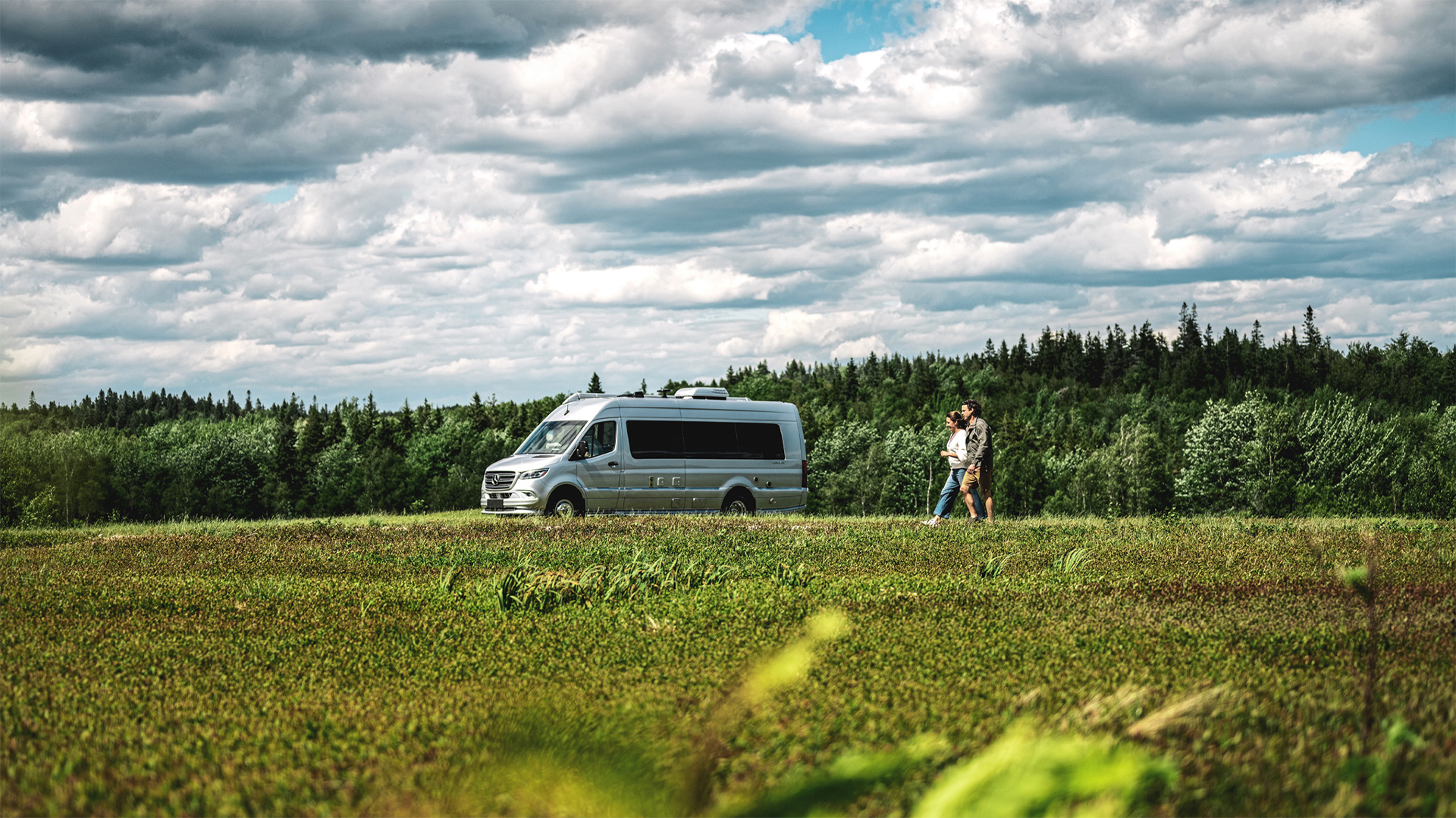 A camper van parked on a lush green field under a partly cloudy sky, with two people walking nearby and a forest in the background.