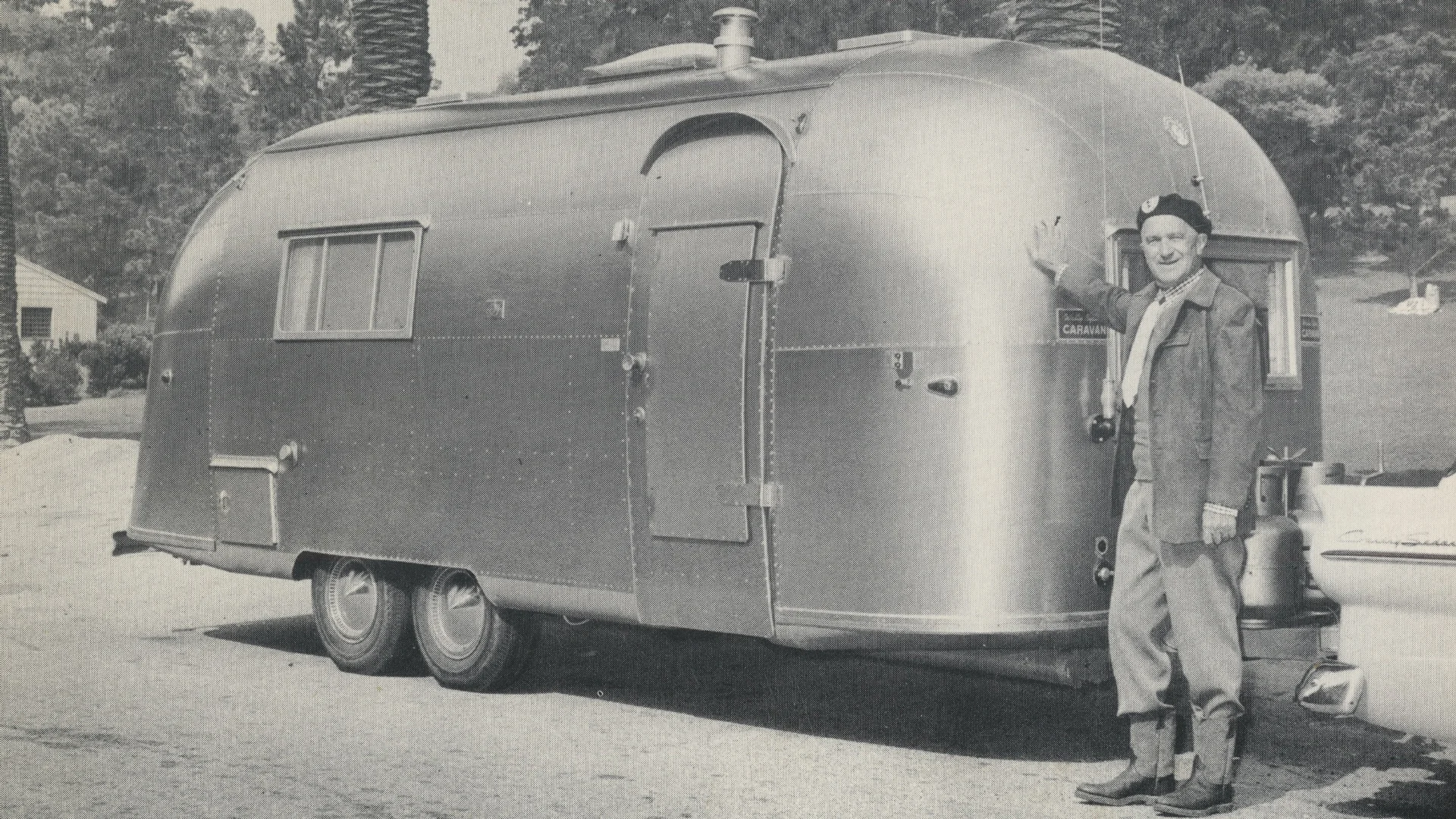 Man wearing a hat and jacket stands beside a vintage Airstream trailer, smiling and touching its side, with trees and houses in the background.