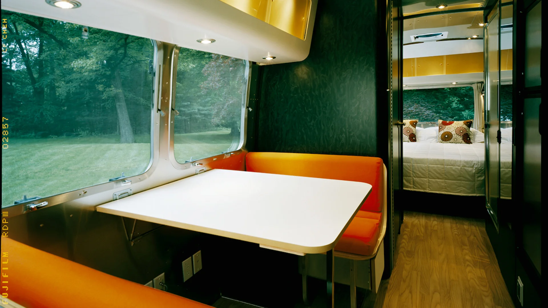 Modern RV interior with an orange and white dining area, large windows with a green outdoor view, and a cozy bedroom in the background.