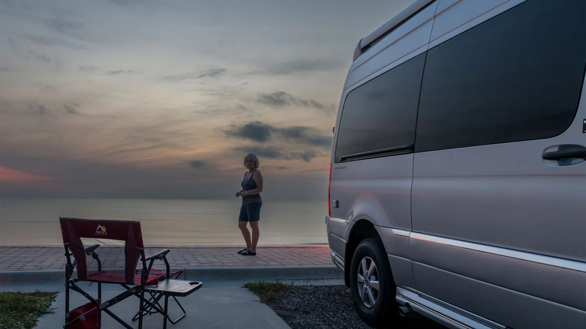 Person standing by the ocean at sunset near a parked van and an empty camping chair.